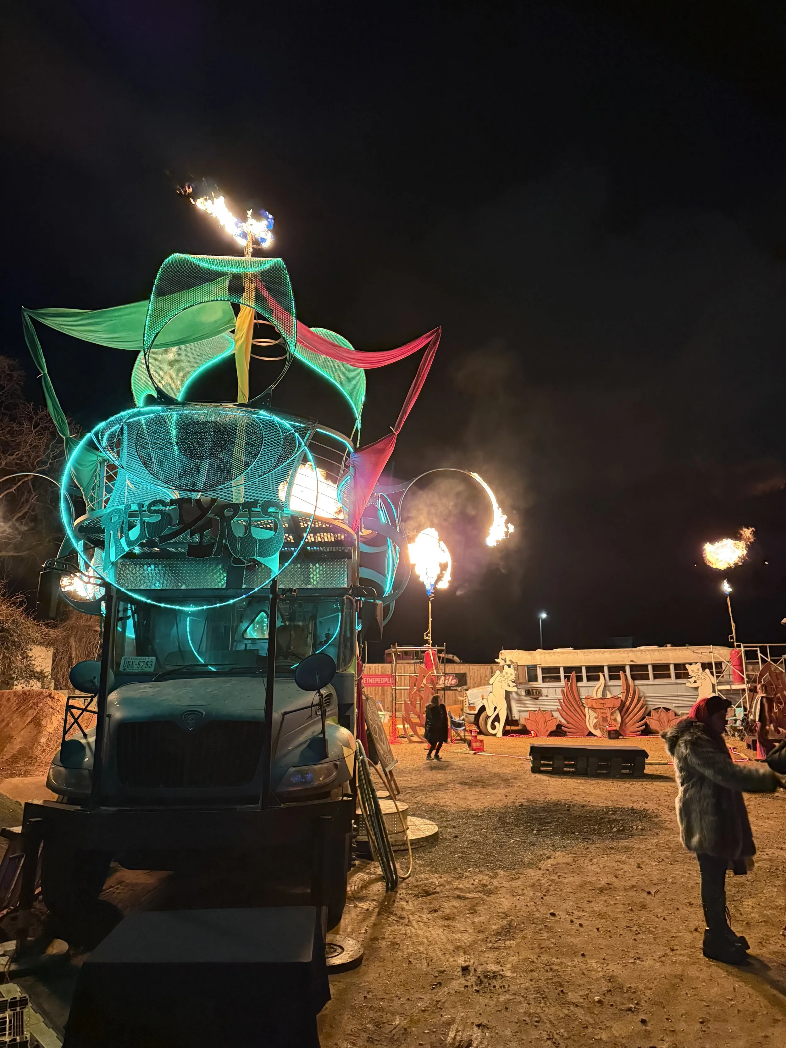 Night scene featuring a decorated truck with neon lights and flames, with a person in a fur coat in the foreground, and a carnival setup with sculptures and more flames in the background.