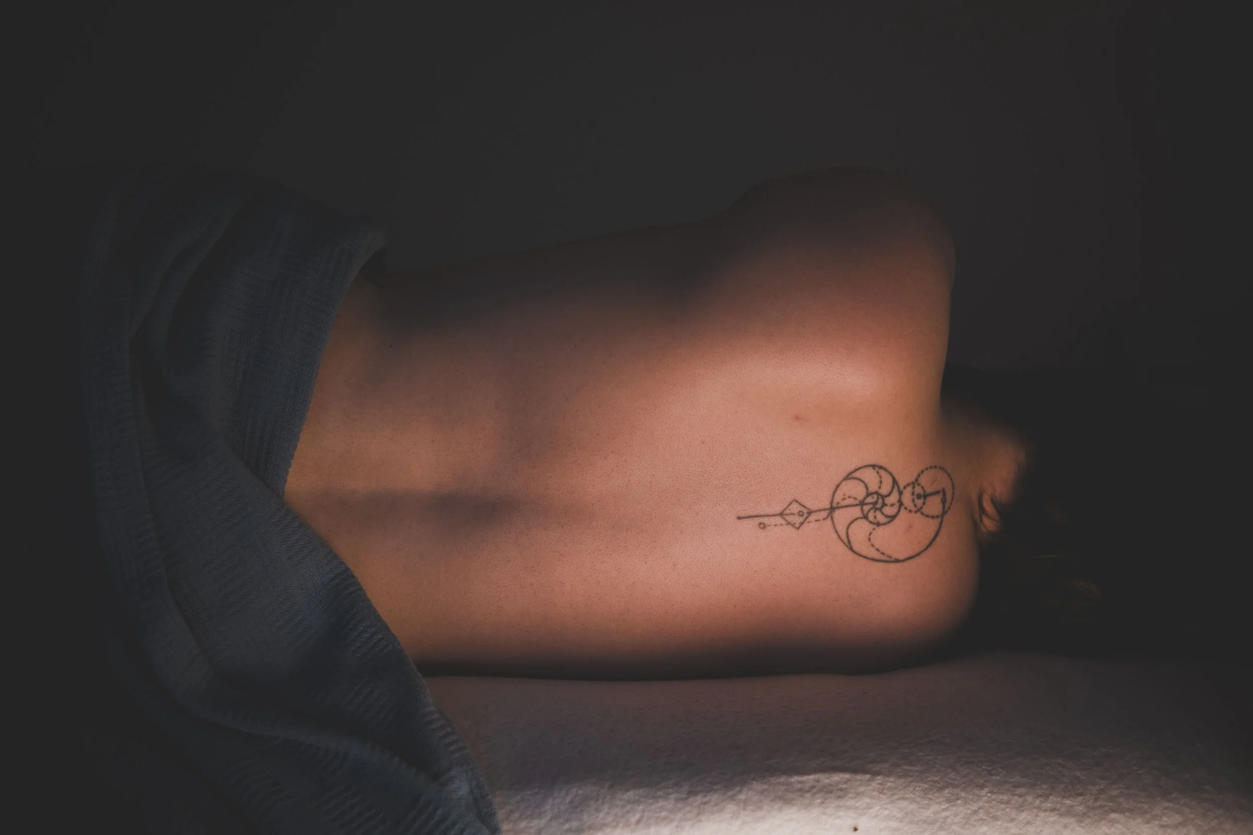 A person lying on their side with a minimalist geometric tattoo on their back, partially covered by a dark-colored blanket or clothing.