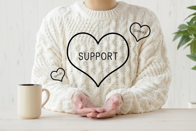 Person wearing a white cable-knit sweater sitting at a table with a beige mug, with three interconnected black hearts containing the word 'SUPPORT' on the sweater.