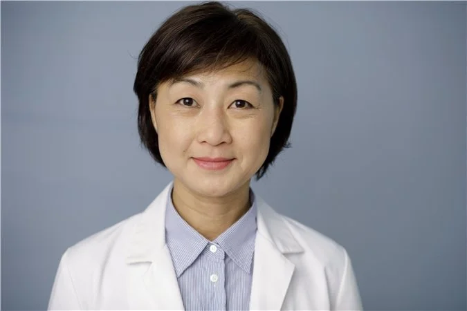 A smiling Asian woman with short black hair, wearing a white lab coat over a light blue collared shirt, posing against a plain gray background.