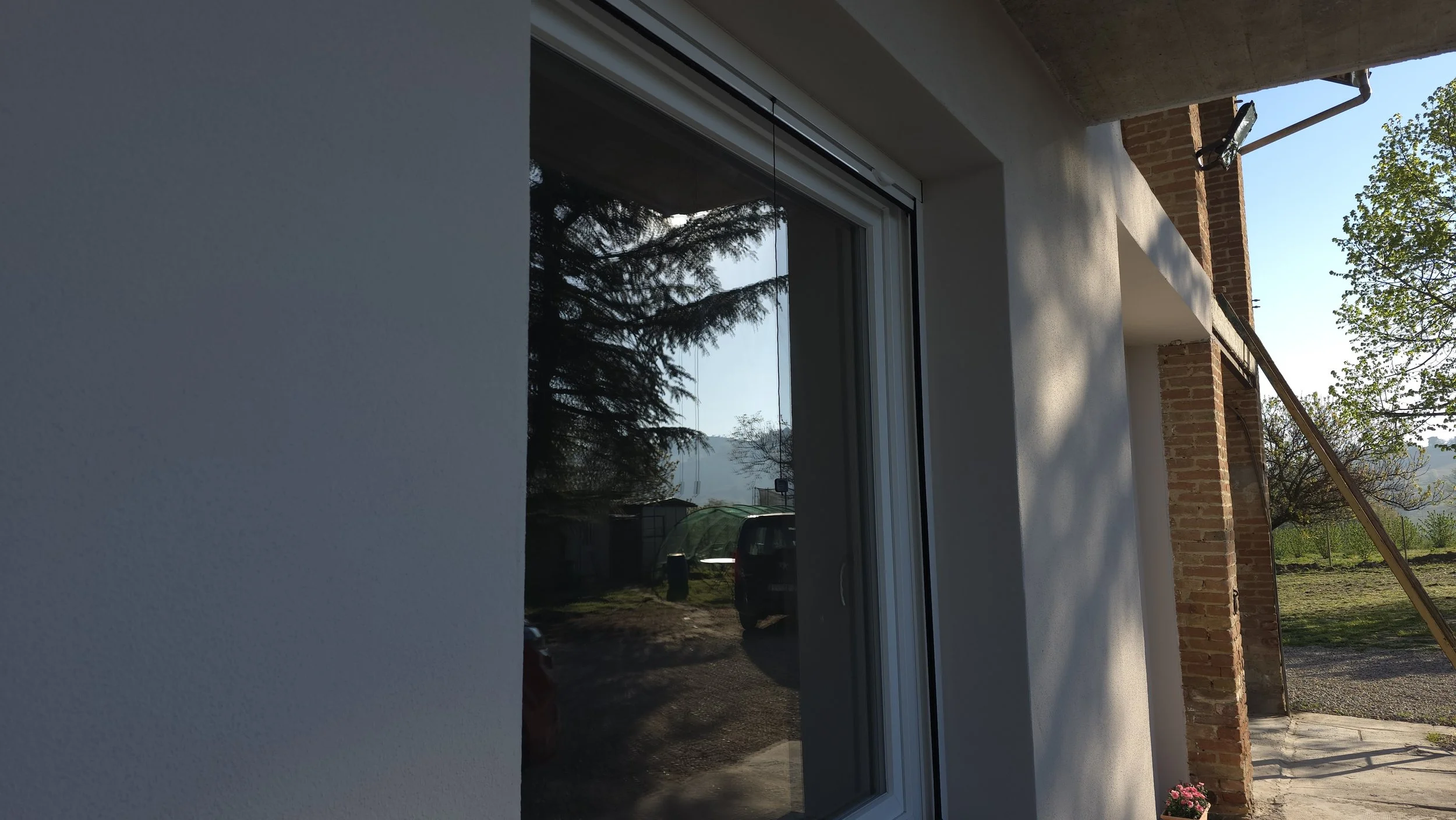 Vista esterna di una parete di una casa con una finestra e parte di un balcone o terrazza, con un muro in mattoni e un tubo di scarico, circondata da alberi e un paesaggio rurale.