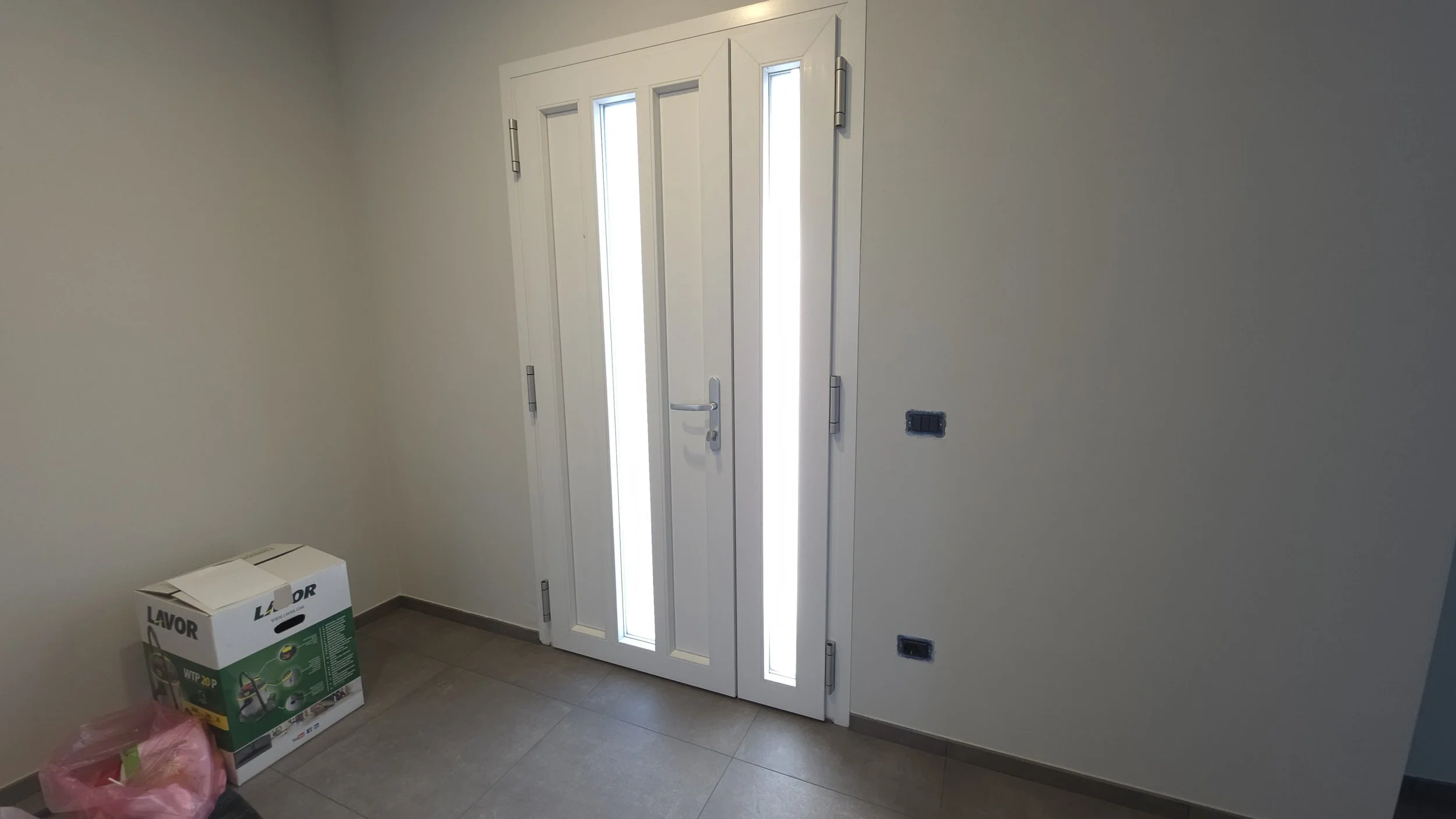 Una porta bianca con vetri laterali e illuminazione naturale. Sul pavimento ci sono scatole di cartone e un sacchetto rosa, in un ambiente interno.