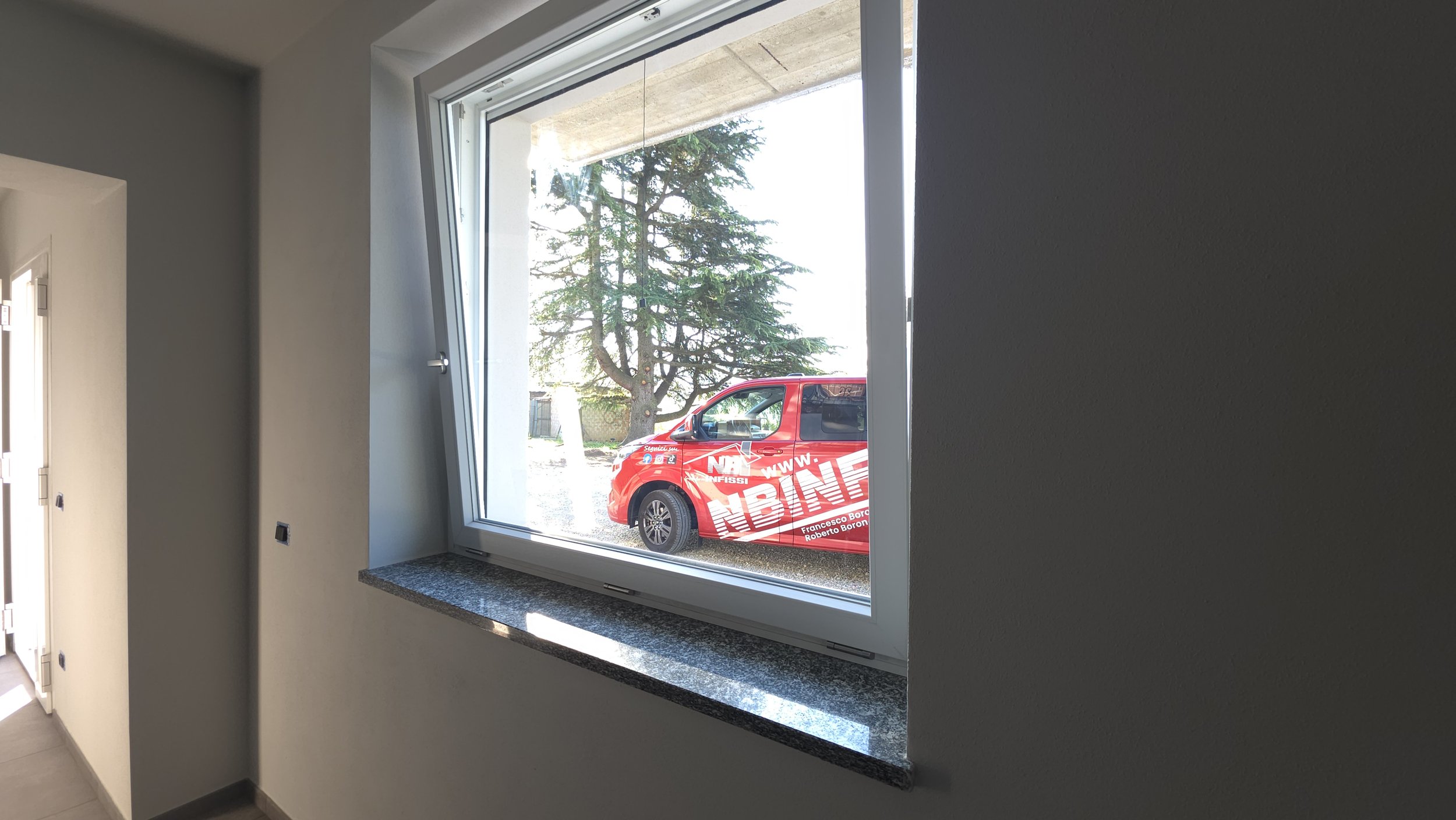 Vista da una finestra in una stanza moderna, con un veicolo rosso parcheggiato fuori, un albero e un cielo chiaro.
