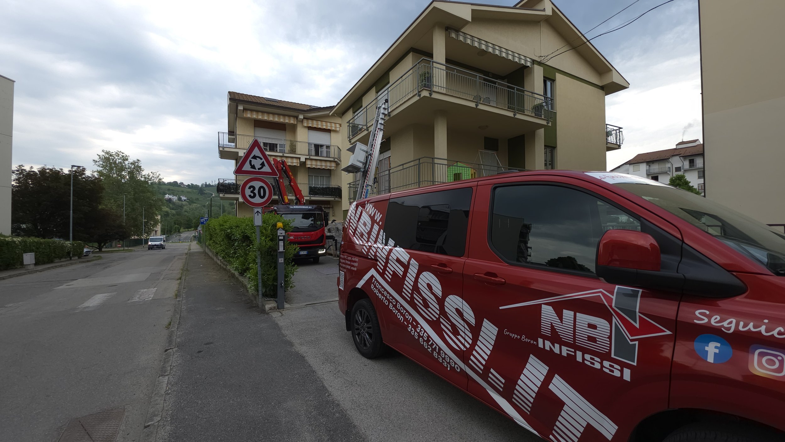 Vettura rossa da lavoro con logo di un'impresa di servizi, parcheggiata davanti a un edificio residenziale, con cannoni sollevatori per lavori in altezza e segnaletica stradale visibile.