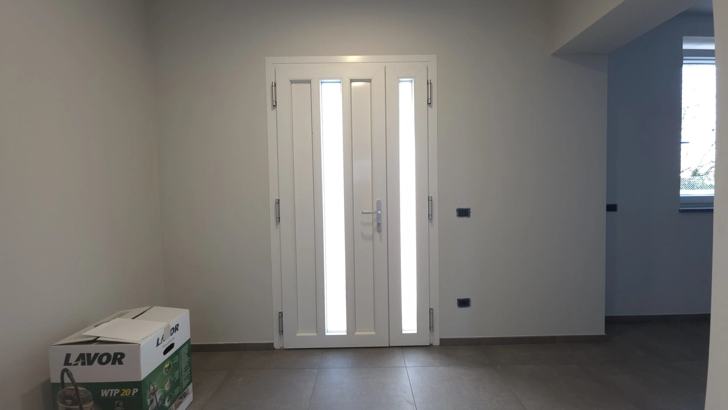 Porta bianca in una stanza luminosa con finestre, con scatole di imballaggio sul pavimento.