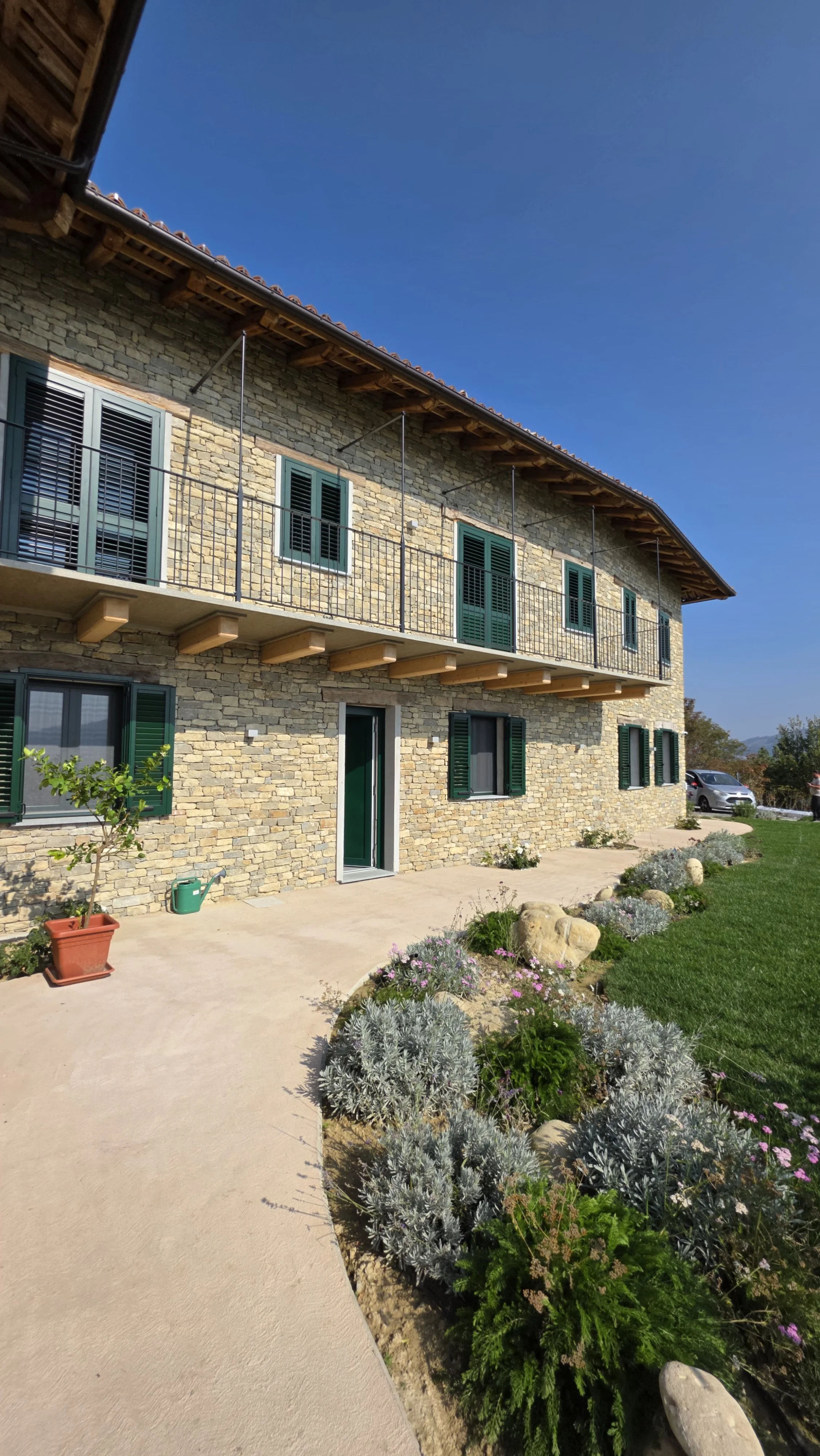 Casa con pietra, finestre con persiane verdi, balcone, giardino con piante e fiori, strada asfaltata