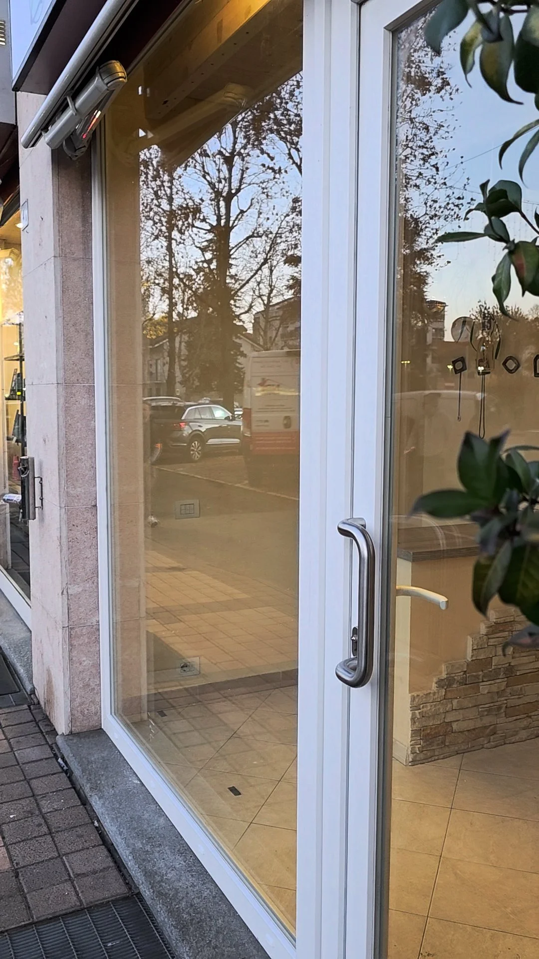 Vetrina di un negozio o ufficio con porta vetrata, riflessi di alberi e auto sulla strada esterna, e interna con parete di mattoni e pavimento in piastrelle