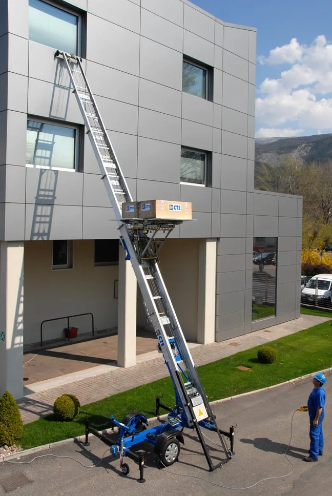 Una macchina per lavori in quota con una lunga scala estensibile, utilizzata da un operatore con casco blu. La macchina è posizionata davanti a un edificio moderno con facciata rivestita in pannelli grigi, e il cielo è sereno con alcune nuvole.