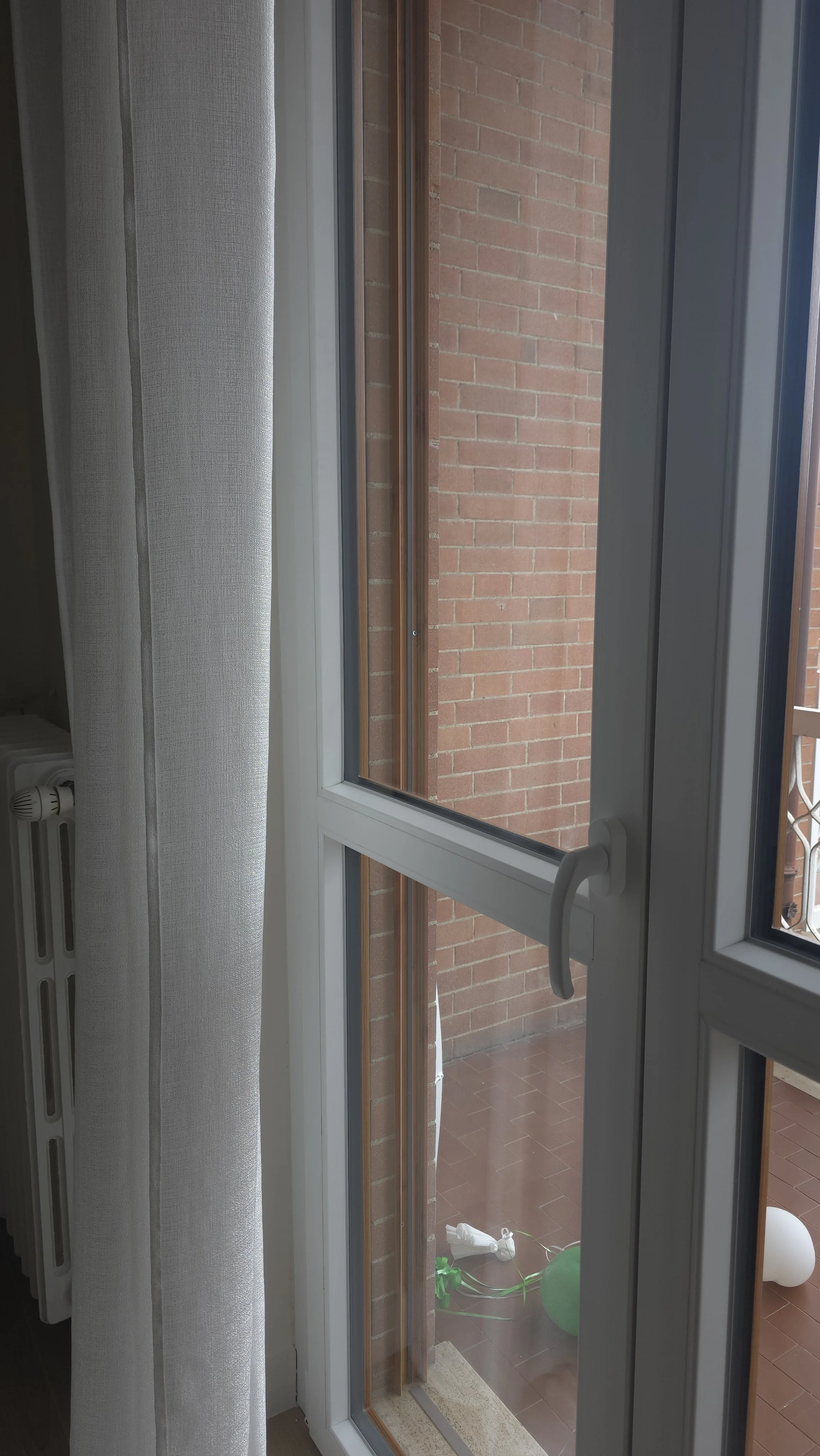 Vista di una porta finestra con vista su un balcone con pavimento in piastrelle, presenti un palloncino verde e un giocattolo bianco in plastica, e sullo sfondo un muro di mattoni.