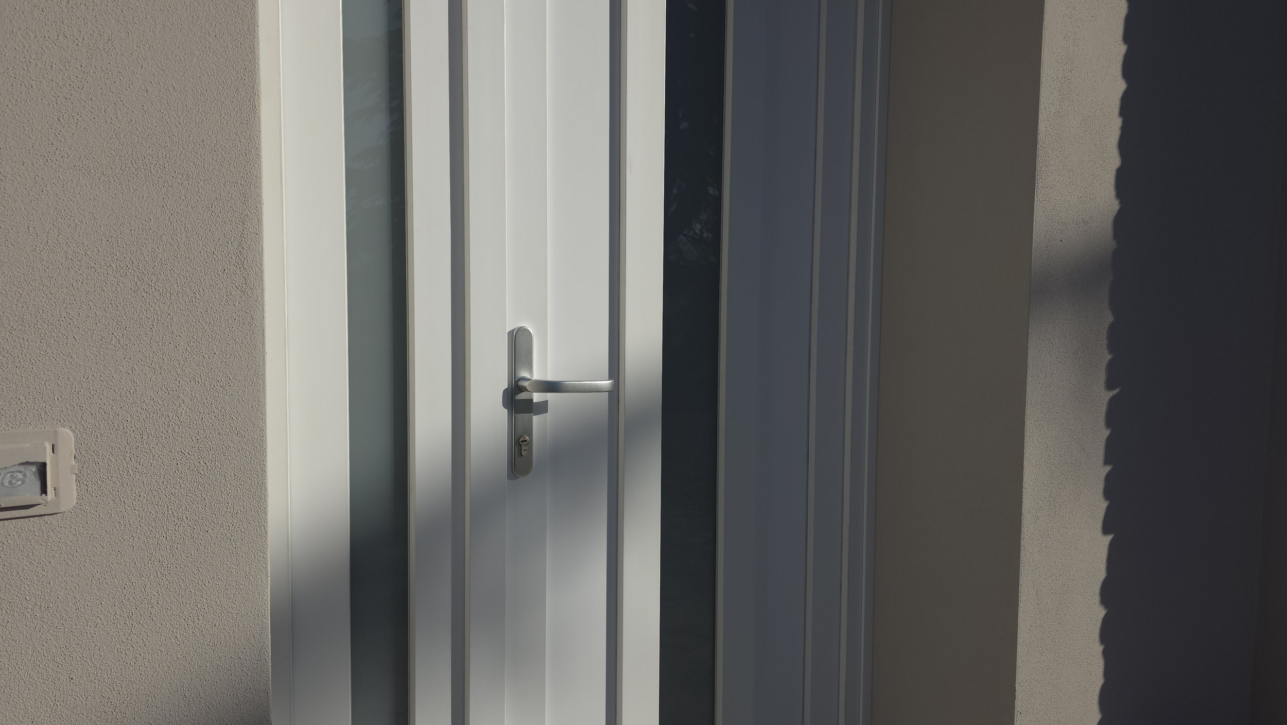 Porta bianca con maniglia e vetri, all'esterno un muro beige e una presa elettrica.