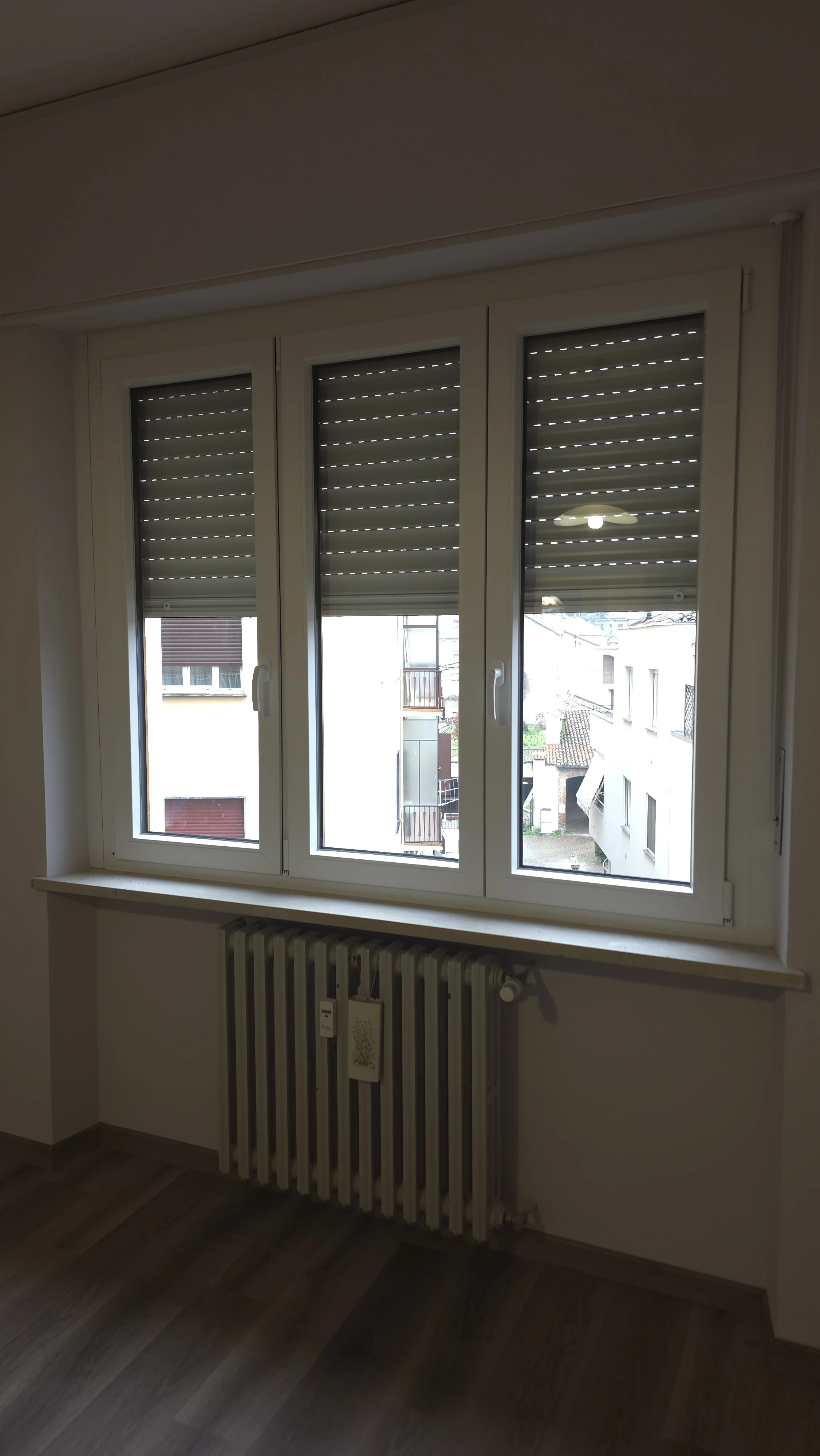 Finestra a doppia anta con tapparelle in posizione semi-abbassata, vista su un cortile con altri edifici.