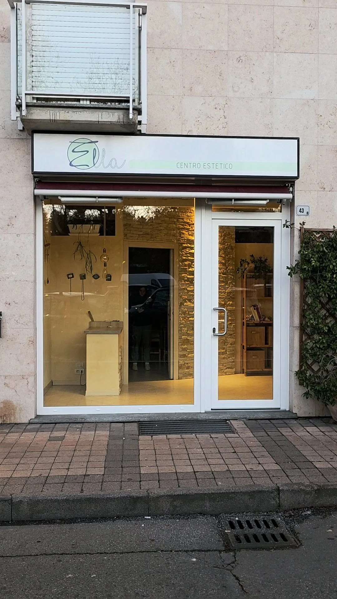 Vetrina di uno studio estetico chiamato Ella Centro Estetico, con porta in vetro e pareti di pietra decorativa interna, all'esterno un marciapiede e una gronda con grate di scarico.