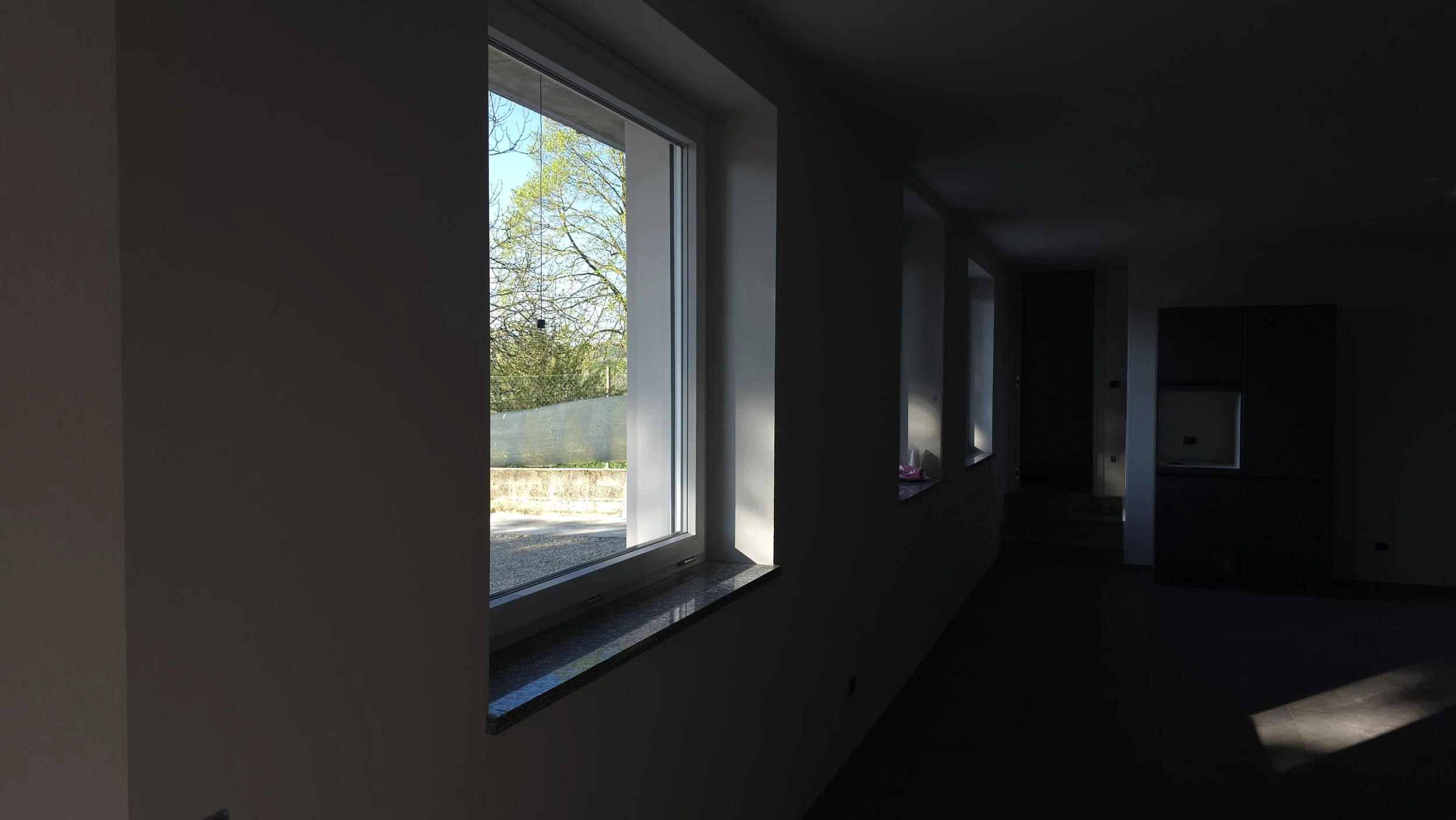 Interno di una stanza con pareti chiare, una grande finestra che lascia entrare luce naturale e mostra alberi verdi all'esterno; arredamento minimale e moderno.