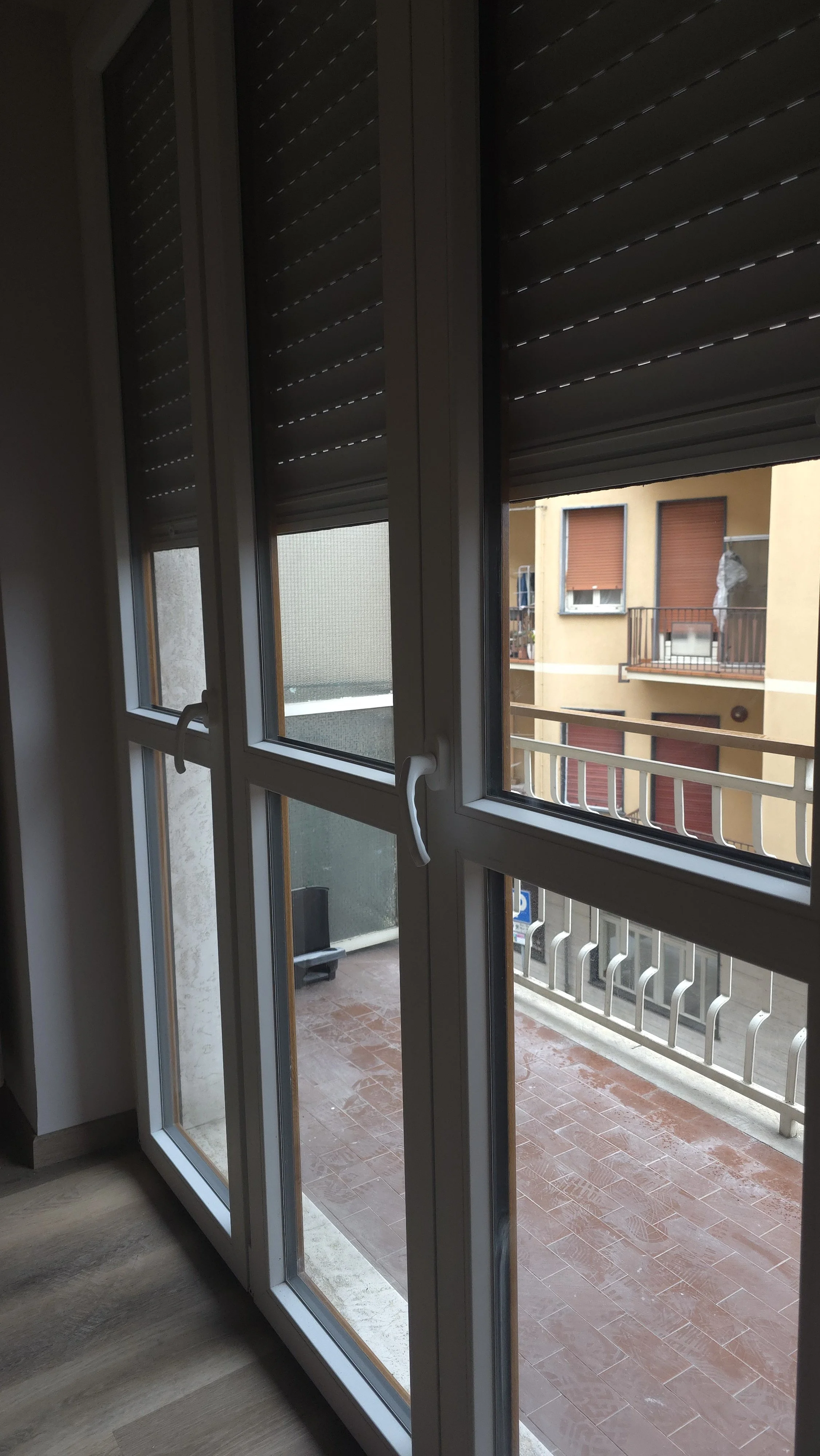 Finestra con tapparelle chiuse che dà su un balcone con pavimento in piastrelle e una ringhiera, con edifici vicini visibili.