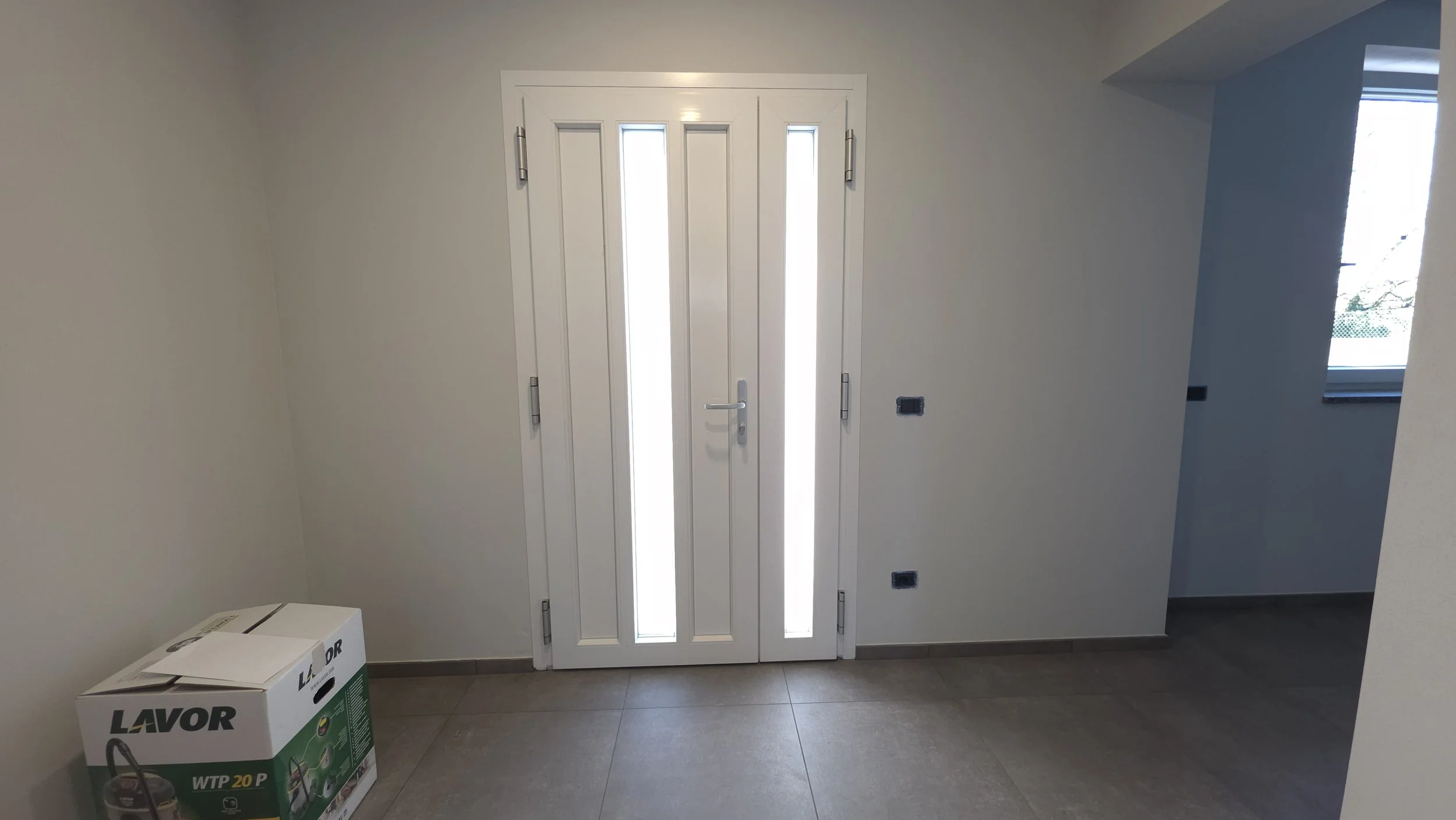Porta bianca con vetri, in una stanza con pareti chiare e una finestra a destra, attualmente in fase di costruzione o ristrutturazione.