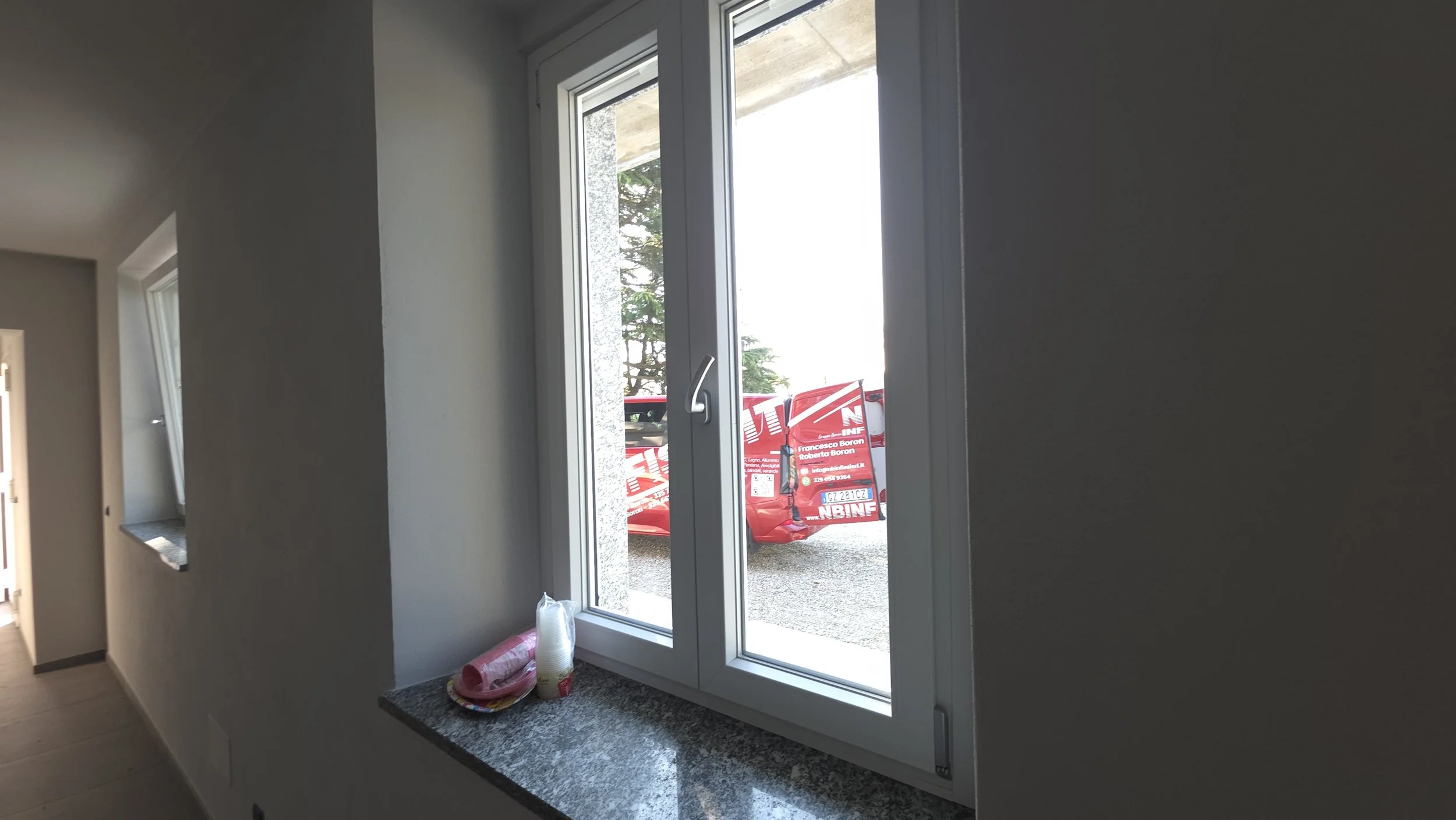 Vista da dentro di una stanza con una finestra e una finestra a doppio battente che si apre all'esterno. Sul davanzale ci sono alcuni rotoli di pellicola protettiva e una pila di bicchieri di carta. Fuori dalla finestra si vede un trattore rosso con 