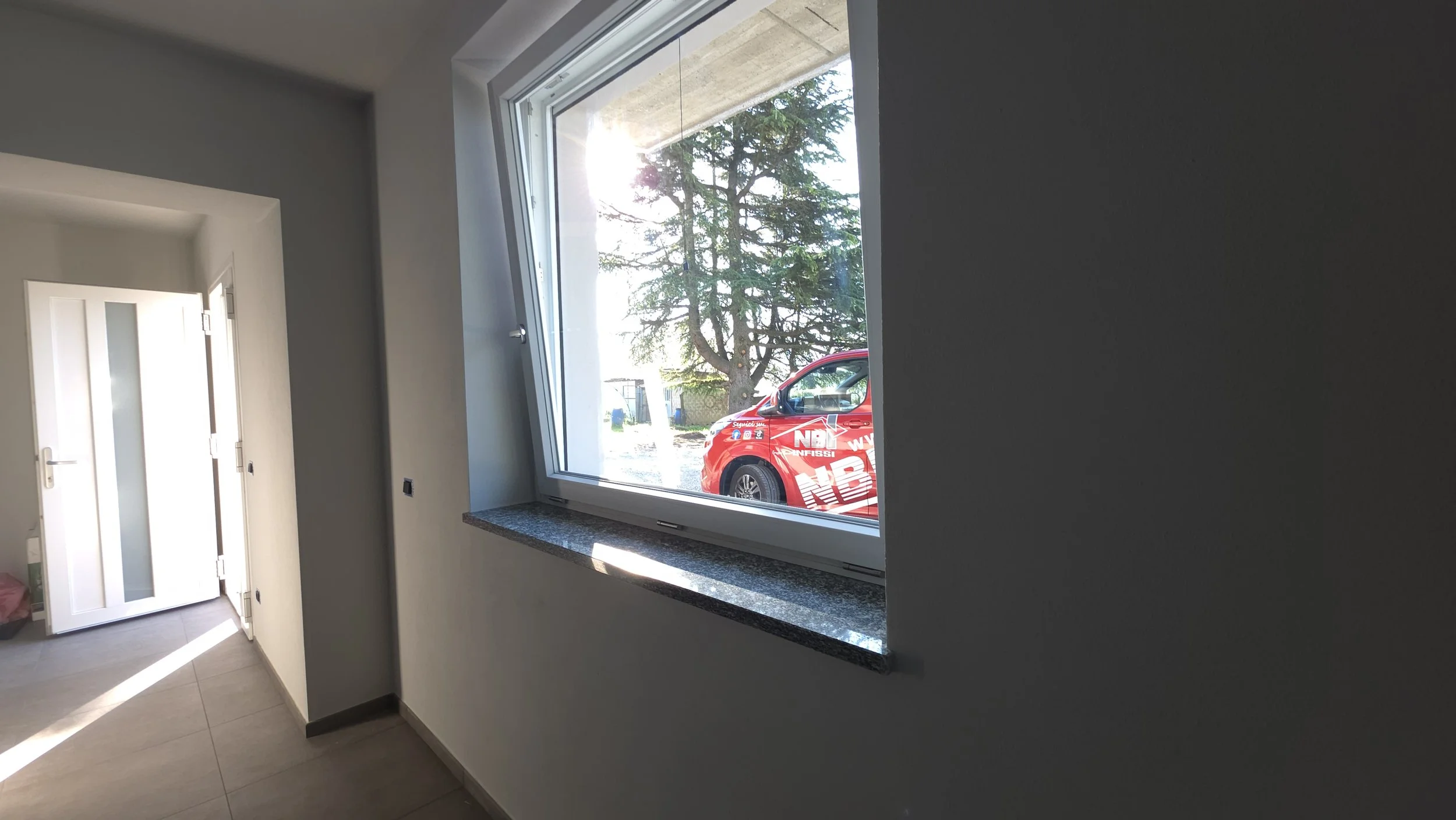 Interno di una casa con una finestra e una porta bianca. La finestra mostra un albero e una macchina rossa con pubblicità parcheggiata fuori. La stanza ha pareti di colore grigio chiaro e pavimento in piastrelle.