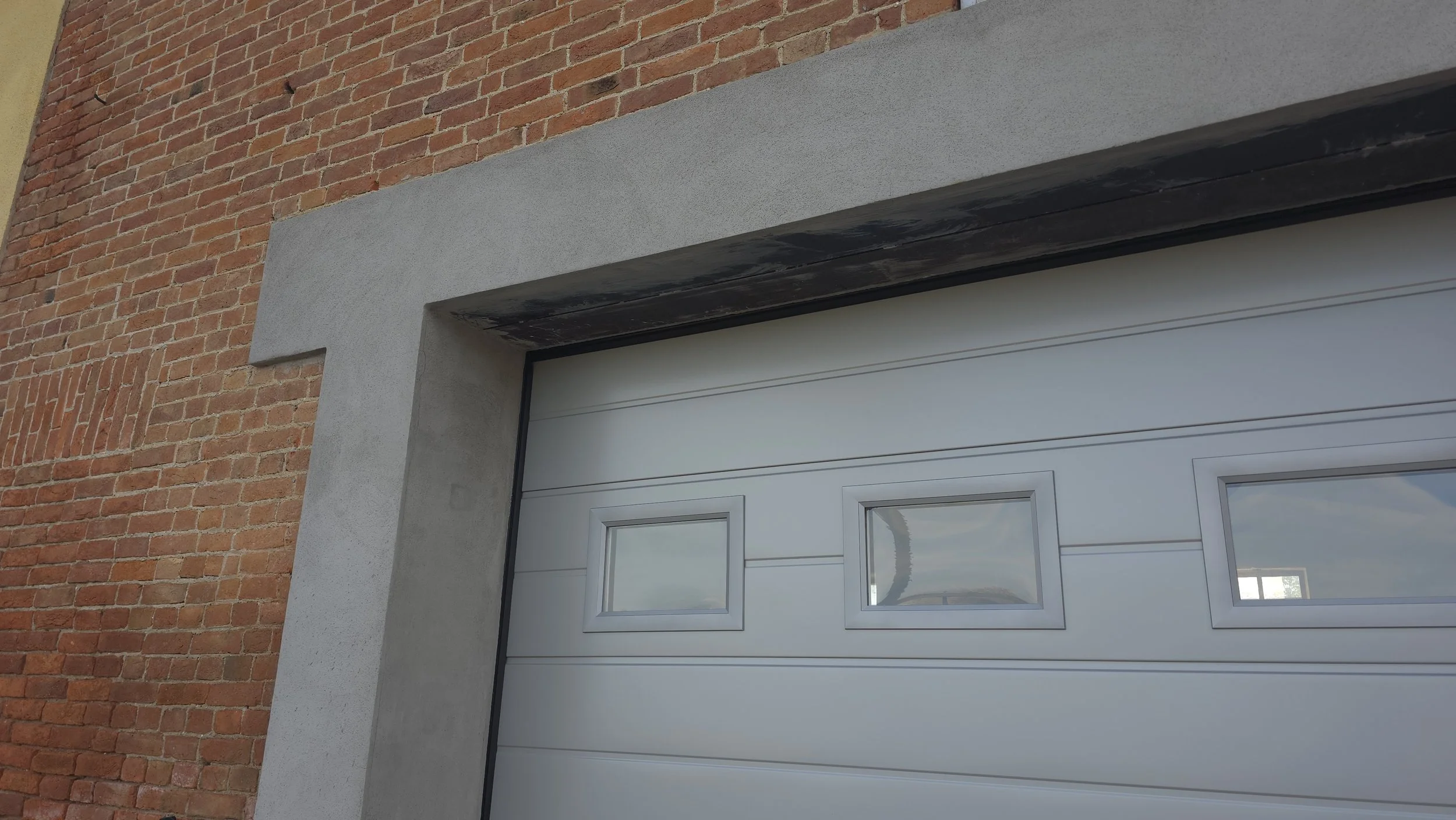 Dettaglio di una porta di garage moderna con finestre, circondata da pareti in mattoni rossi e rivestimento in cemento grigio.