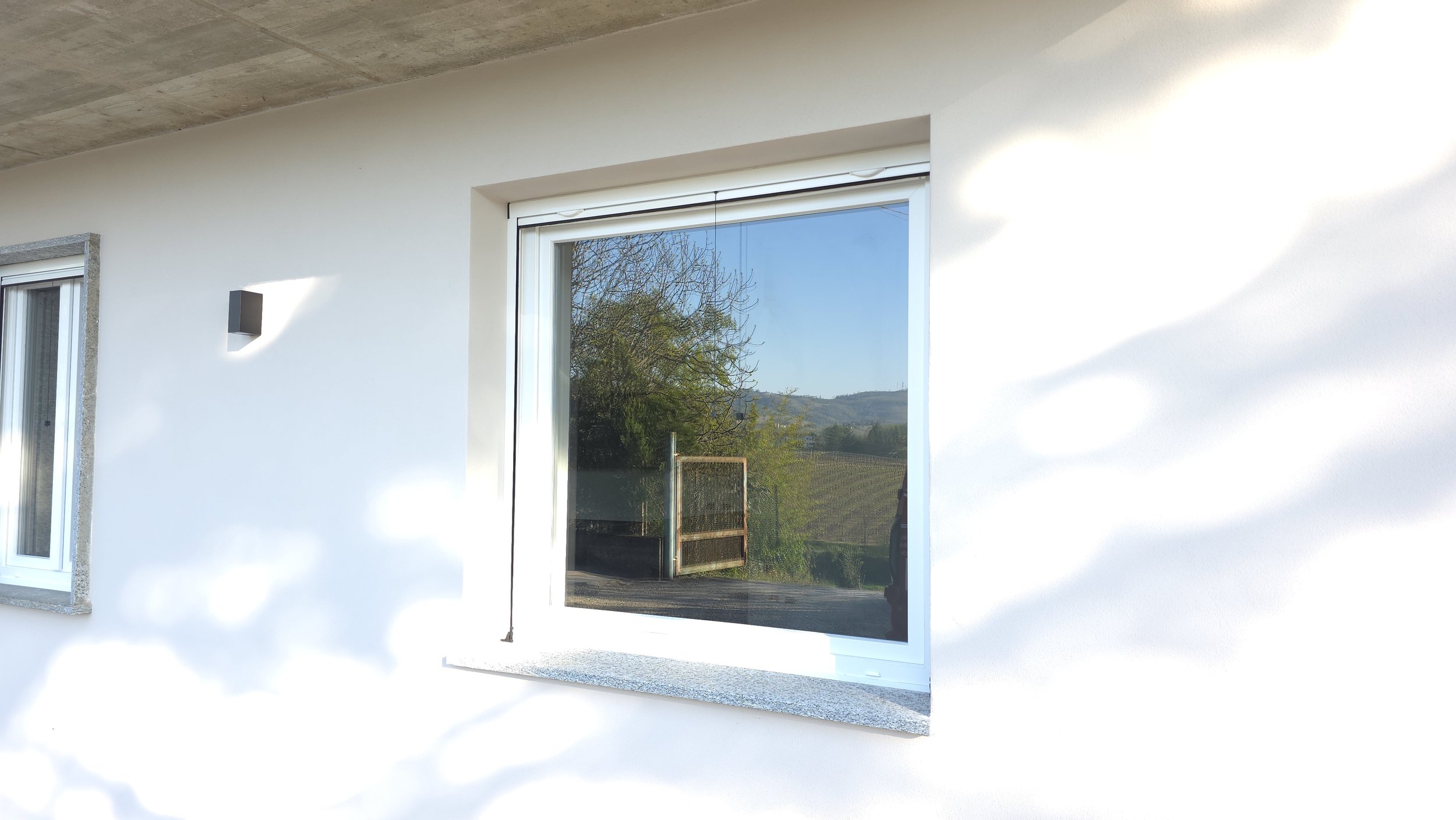 Finestra di una casa con vista su campagna e colline, muro bianco e luce solare.