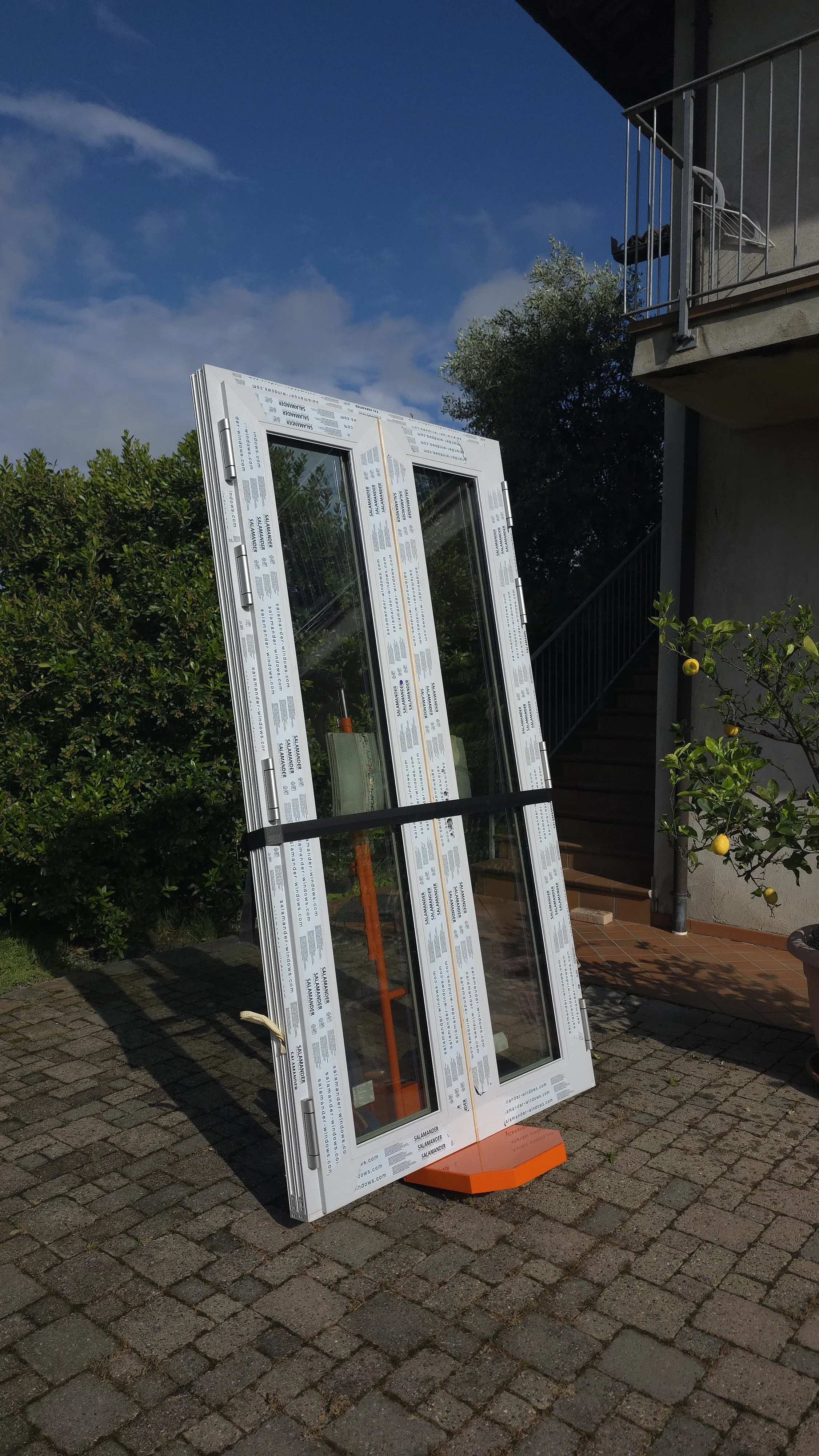 Una finestra in PVC con vetri singoli appoggiata all'aperto su una superficie pavimentata, con un albero di limoni e un'area con scala e balcone sullo sfondo, sotto un cielo azzurro con alcune nuvole.