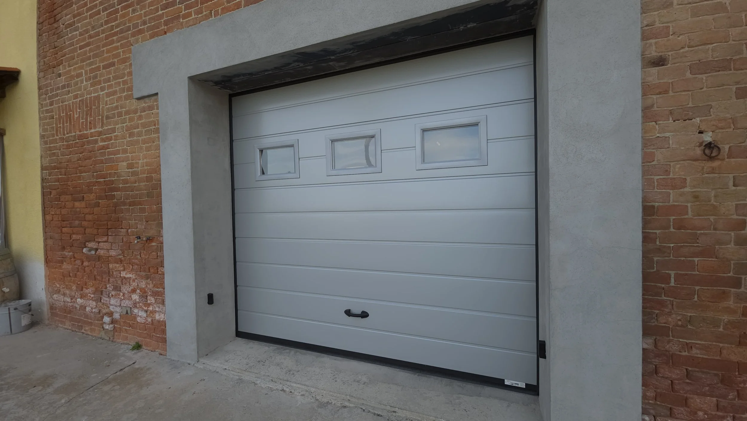 Porta di garage bianca con tre finestre rettangolari, incastonata tra muri di mattoni rossi e cemento.