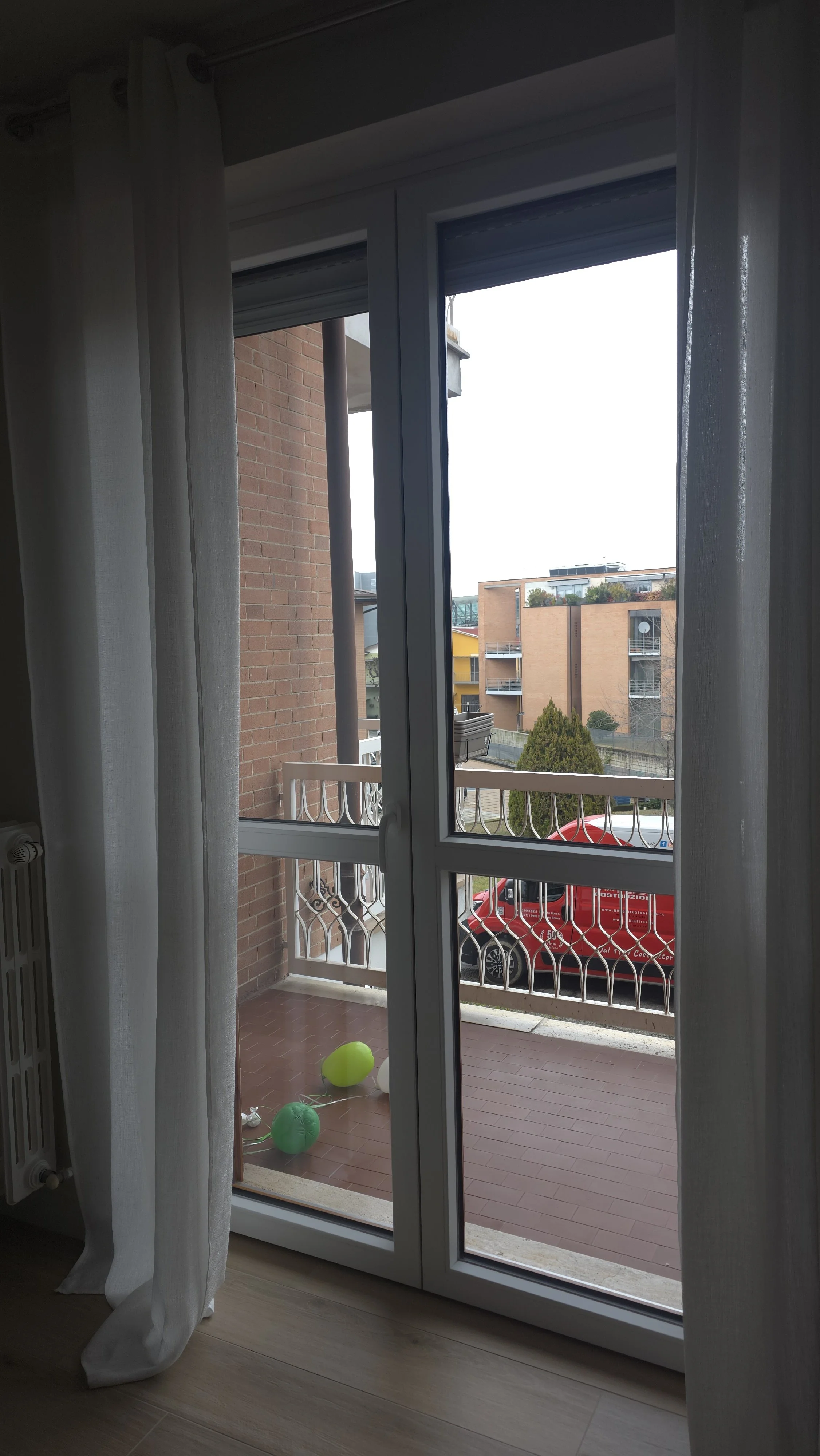 Vista da una finestra sulla terrazza con palloncini colorati a terra e un'auto rossa parcheggiata davanti organizzata sulla strada, edifici residenziali sullo sfondo.