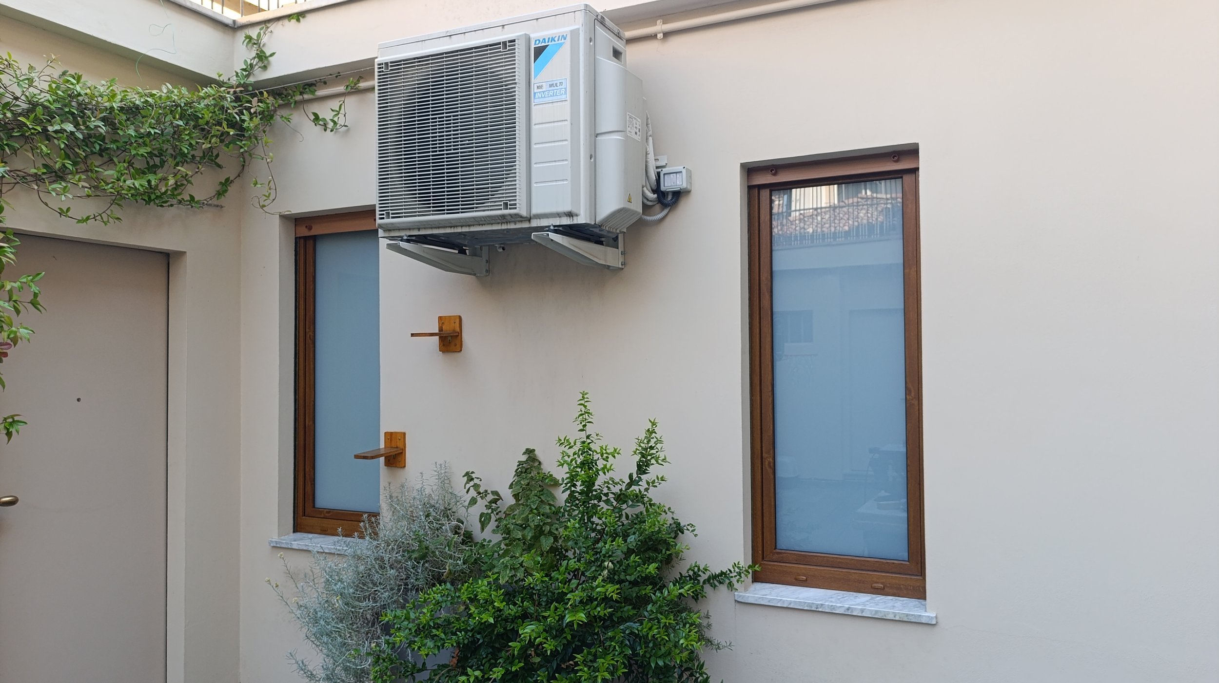 Muro esterno di una casa con due finestre con infissi in legno e un'unità esterna di condizionamento d'aria sopra una delle finestre. Ci sono piante verdi e un'ulteriore finestra a sinistra.