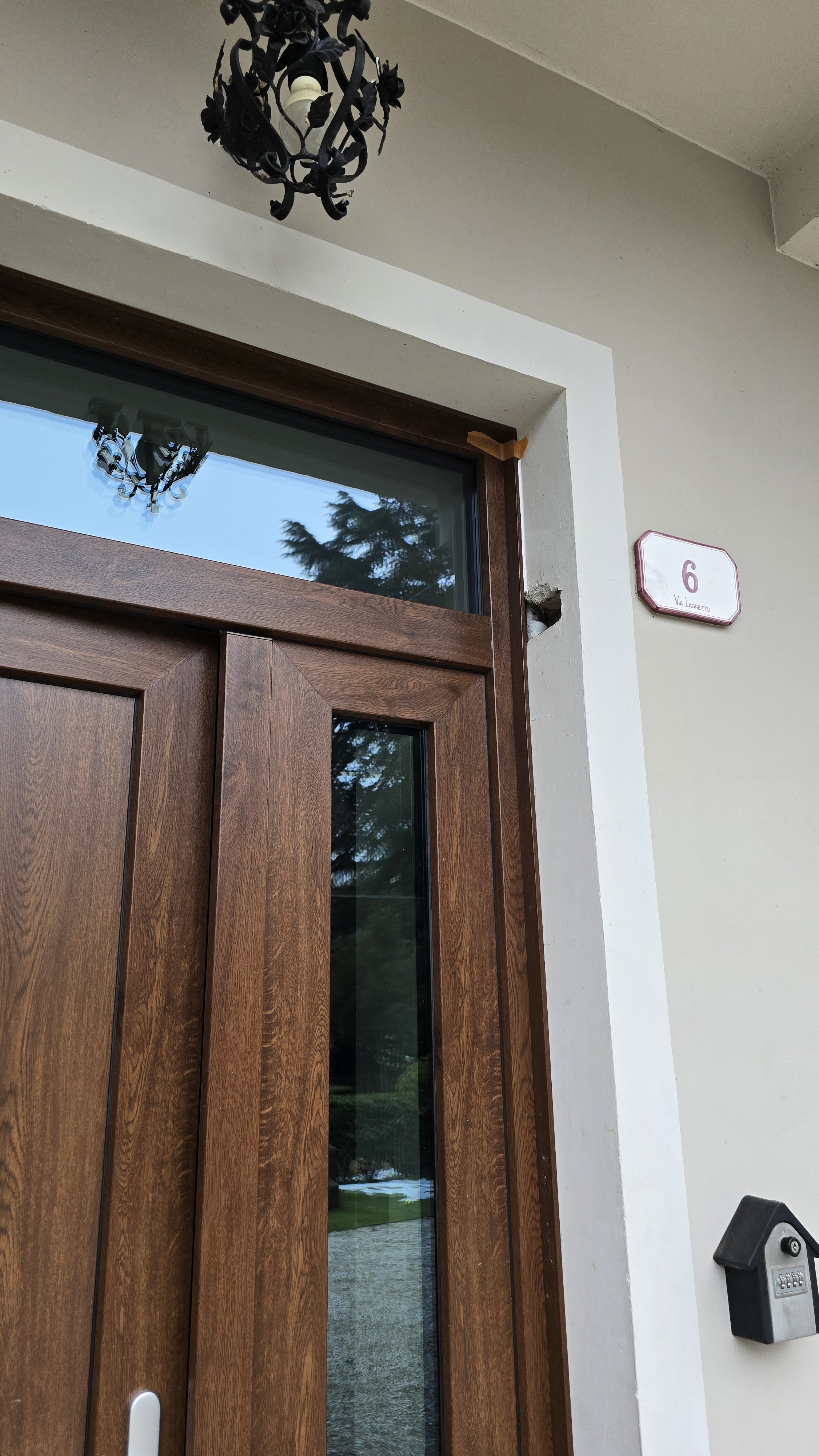 Porta di ingresso in legno con finestra sopra, numero 6 sulla parete vicino, lampadario nero appeso sopra, ed elemente di arredo esterno come la cassetta delle lettere