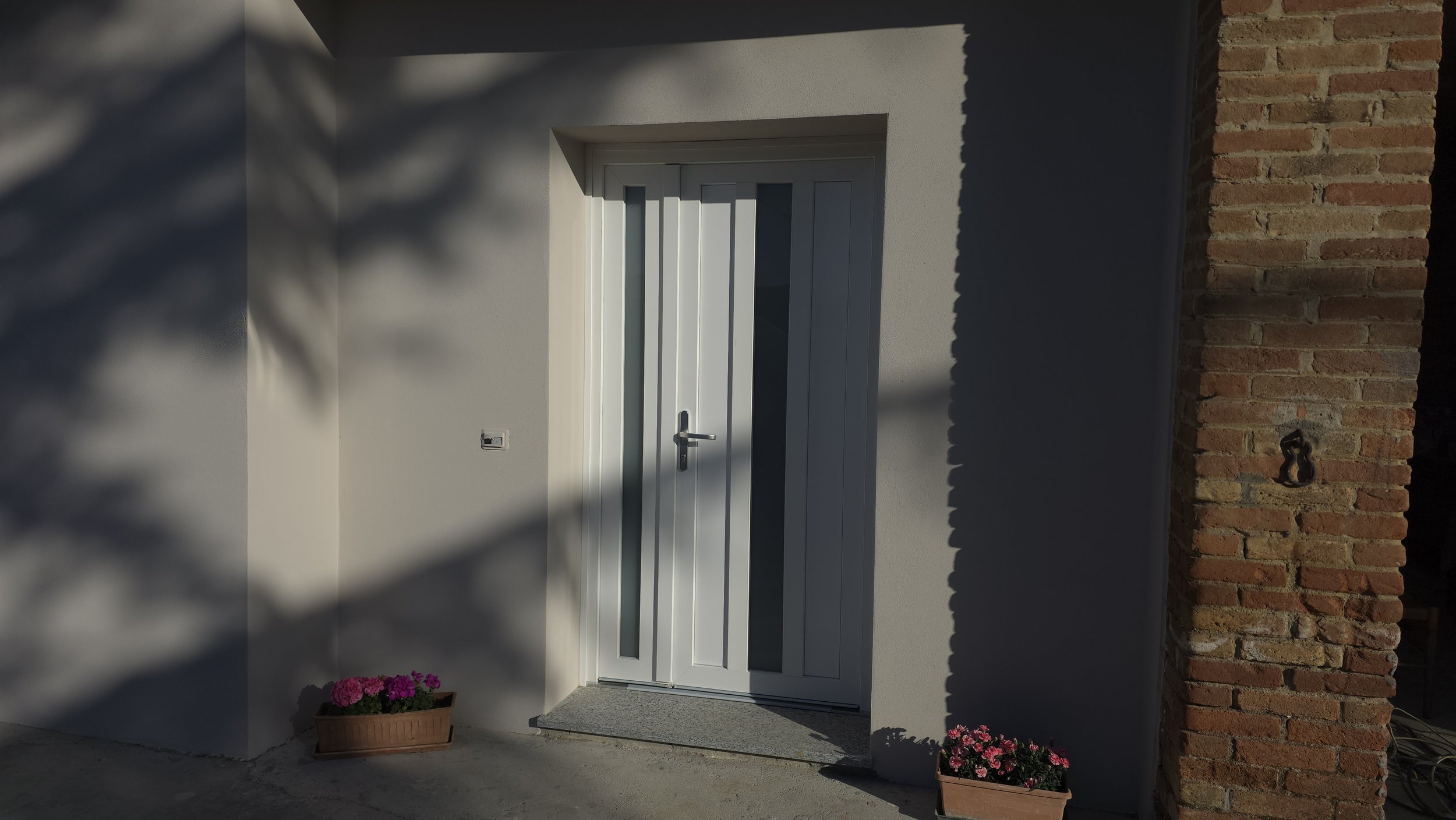 Foto di una porta bianca moderna con vetro, circondata da un muro bianco e una parete di mattoni rossi. C'è una fioriera con fiori rosa alla base della porta, e una piccola scatola di metallo con anello di ferro sulla parete di mattoni.