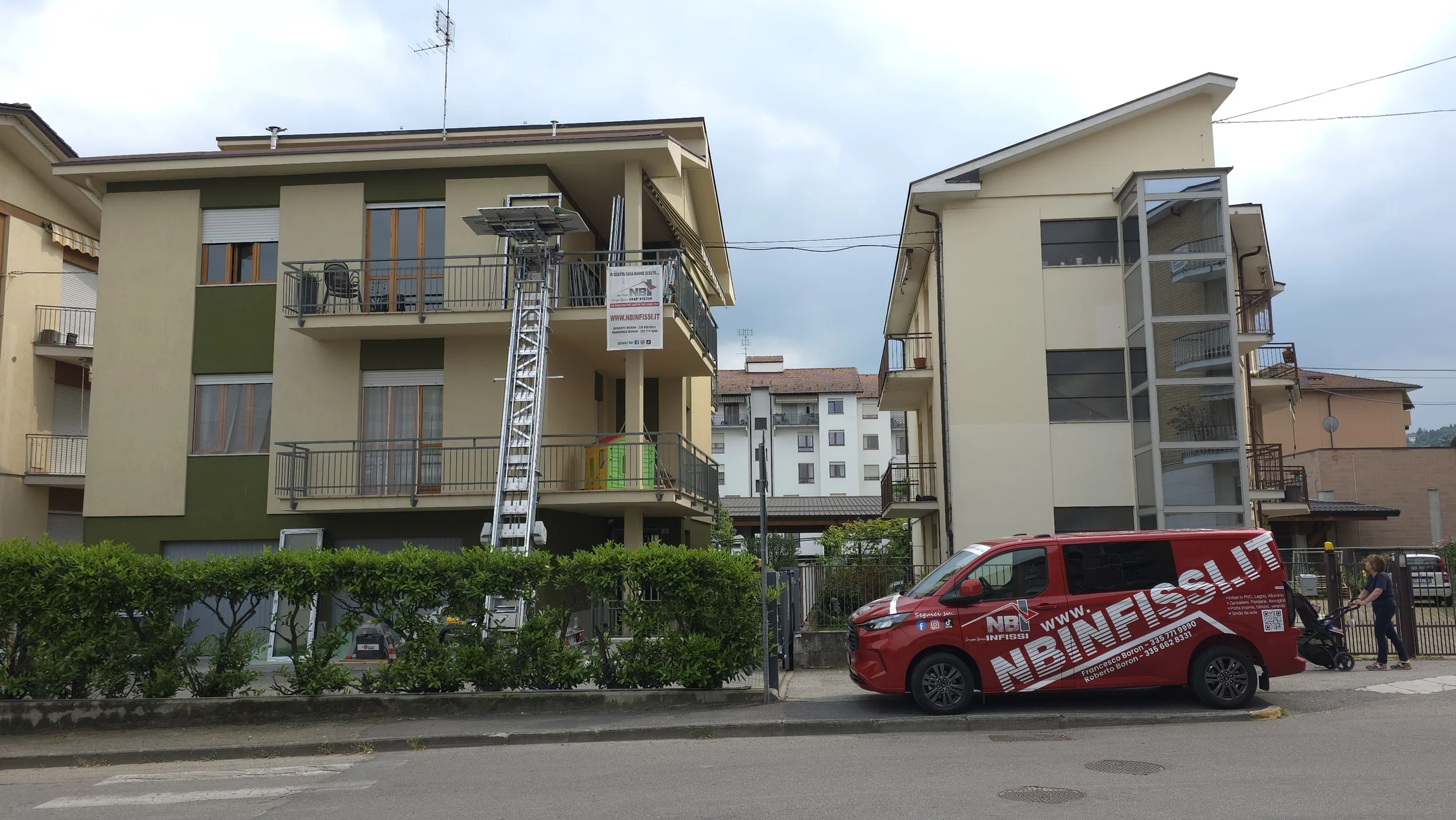 Edificio residenziale a più piani con balconi e finestre. Una scala esterna è coperta da una struttura in vetro. Davanti, un'auto rossa con pubblicità della ditta NB InFissi e un uomo con una bicicletta.