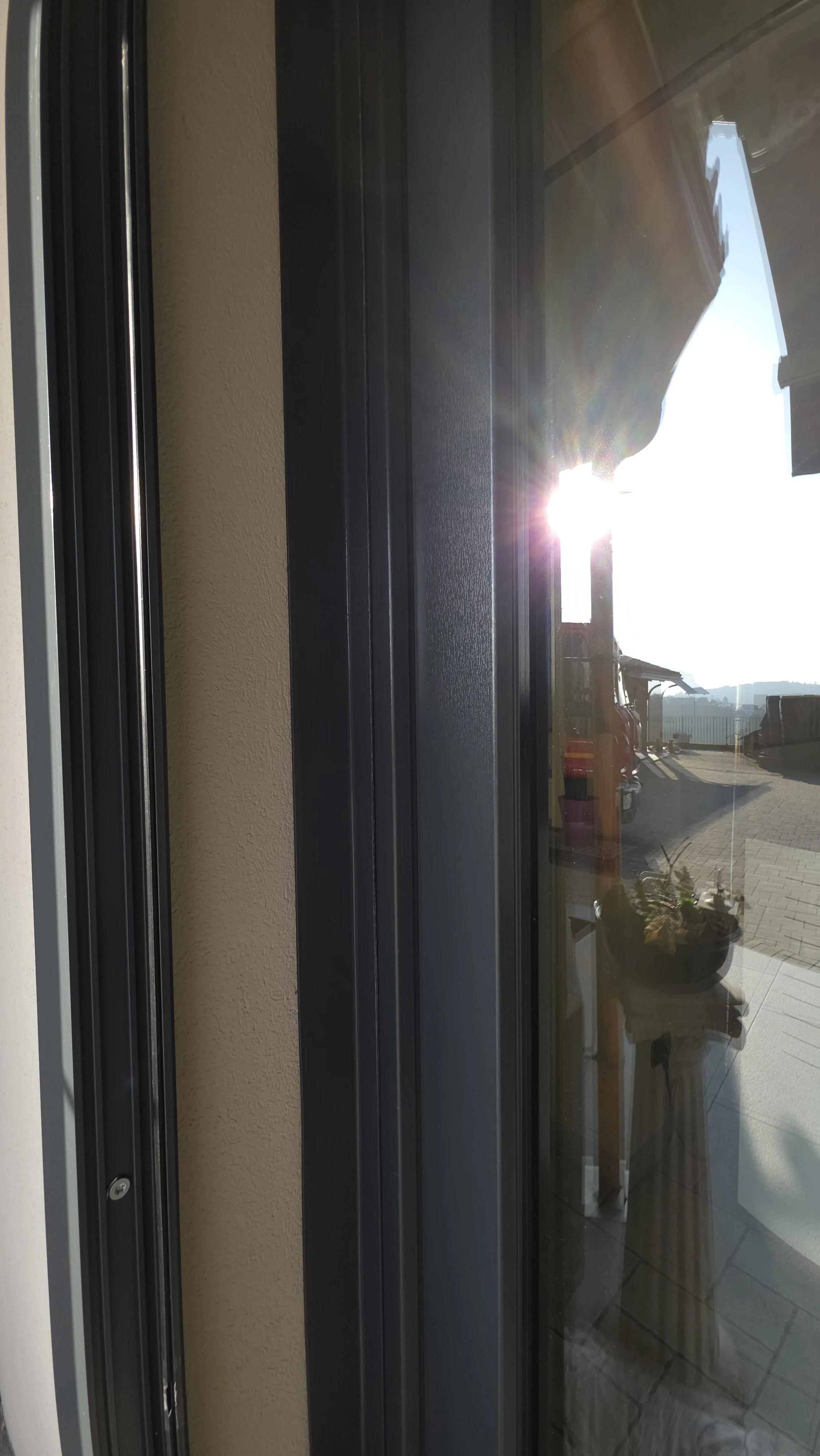 La foto mostra una finestra con il sole che splende e riflette sul vetro, creando un bagliore luminoso, con un arredamento e un balcone visibili attraverso il vetro.