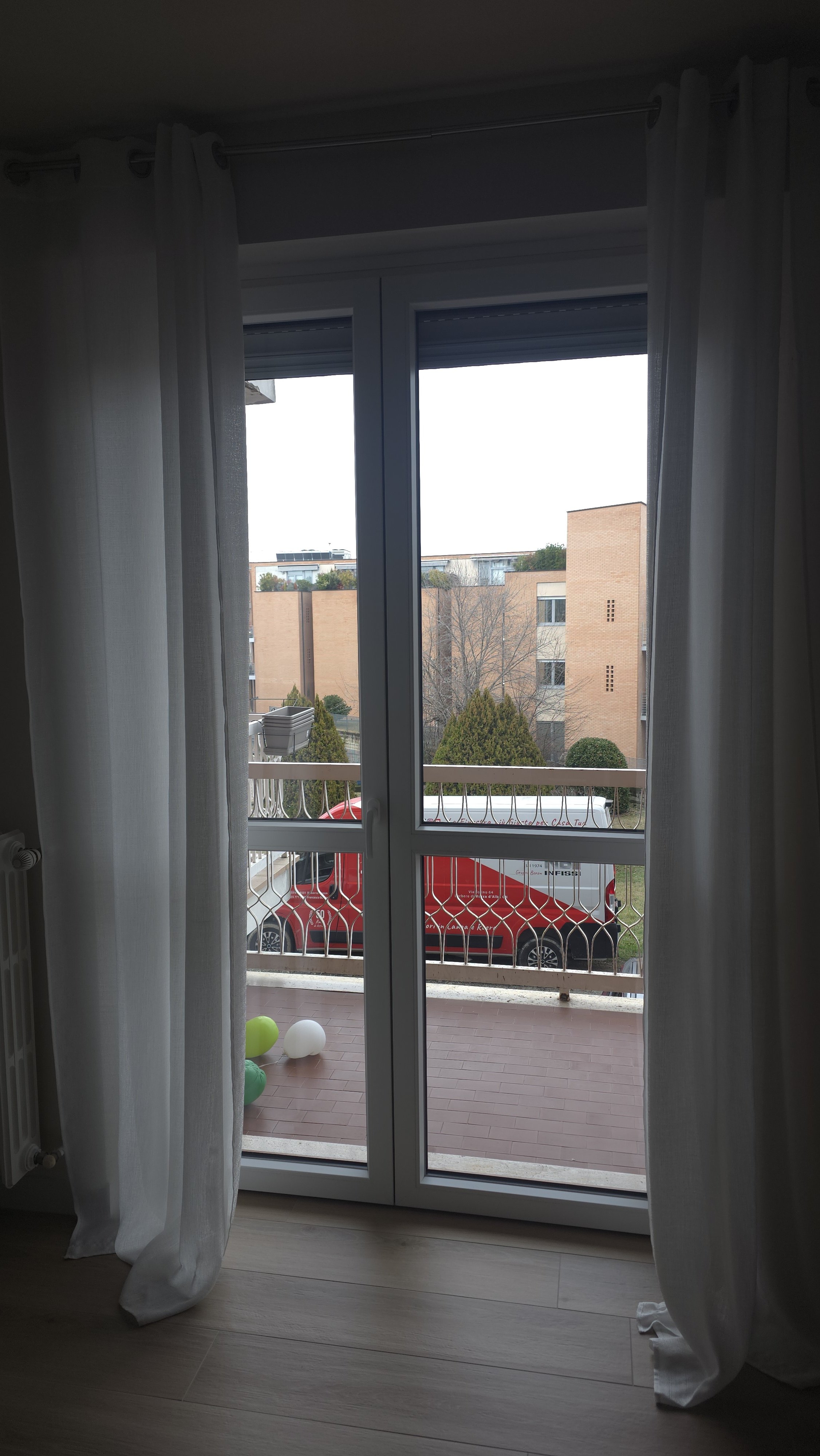 Vista attraverso una porta con vetri su un balcone con palloncini e un veicolo rosso parcheggiato fuori.