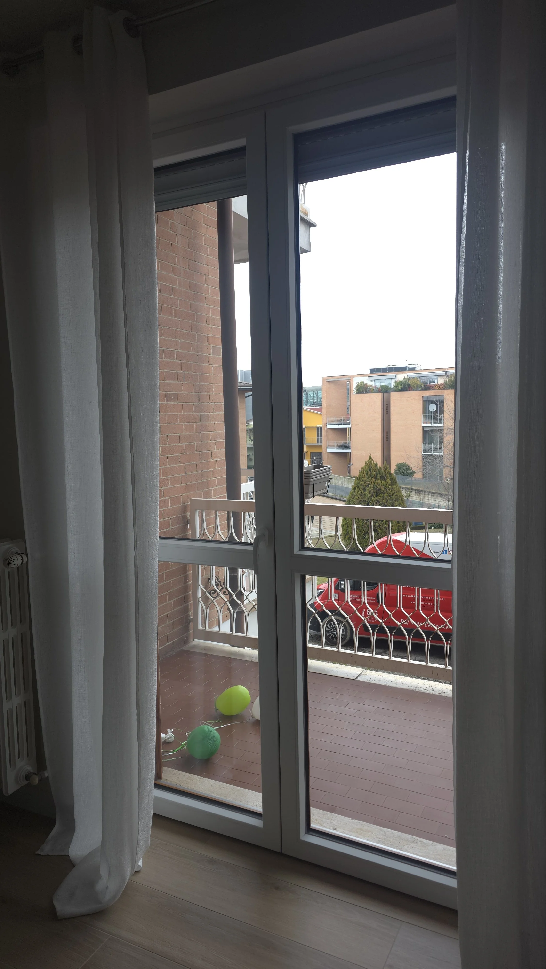 Vista da una finestra su un balcone con palloni gonfiabili di vari colori, un'auto rossa parcheggiata, edifici di appartamenti e un albero.