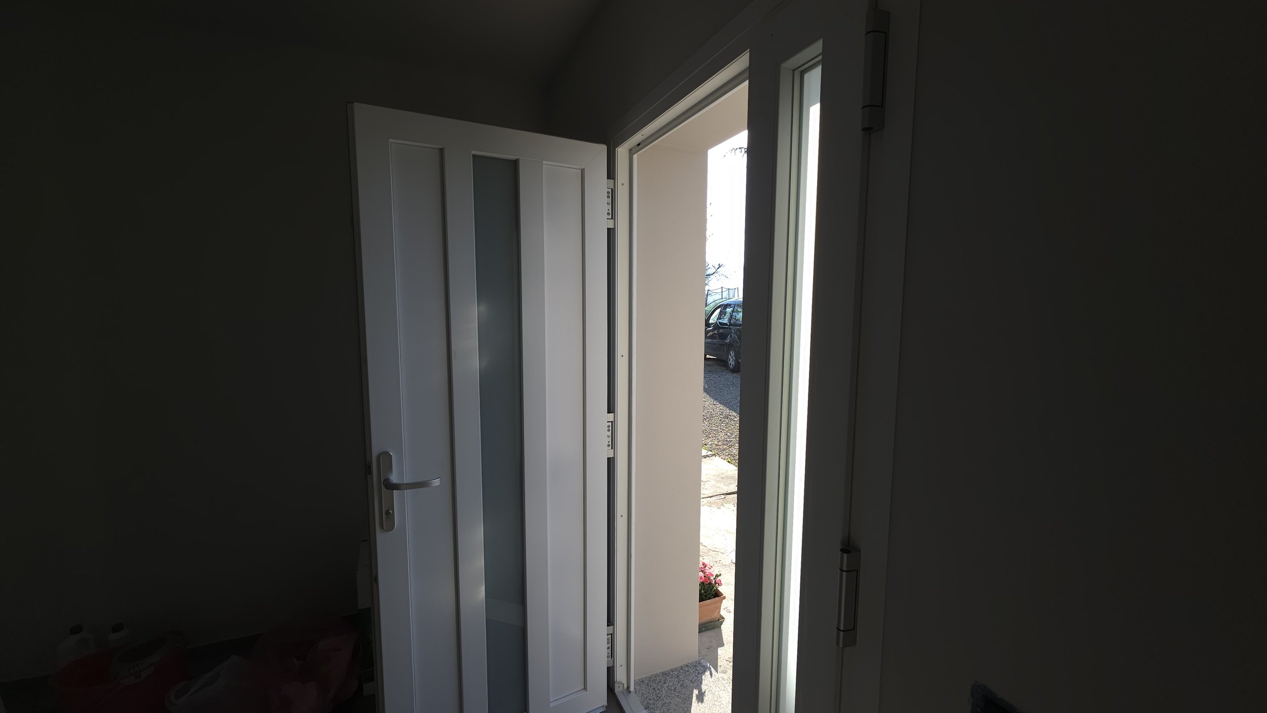 Ingresso di casa con porta bianca e finestra, vista esterna con veicoli e piante