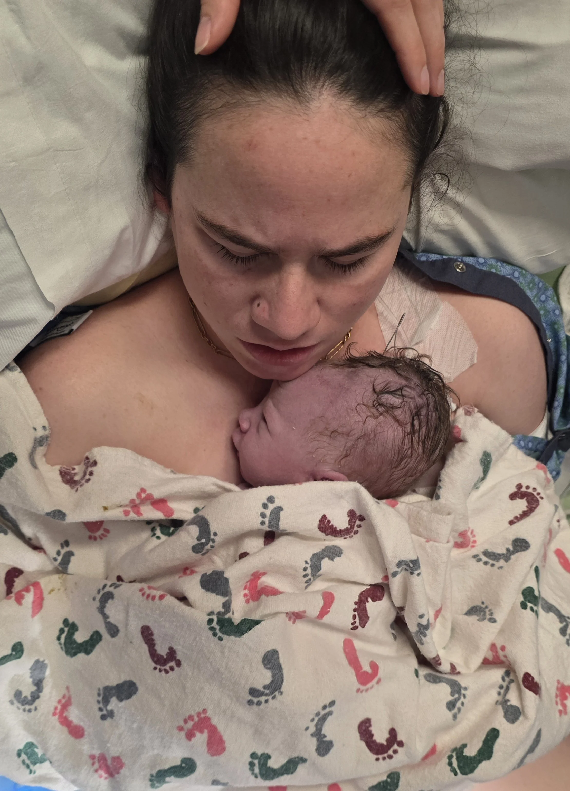 A woman lying in a hospital bed holding a newborn baby close to her chest, both wrapped in a blanket with colorful footprints pattern, in a moment of bonding and tenderness.
