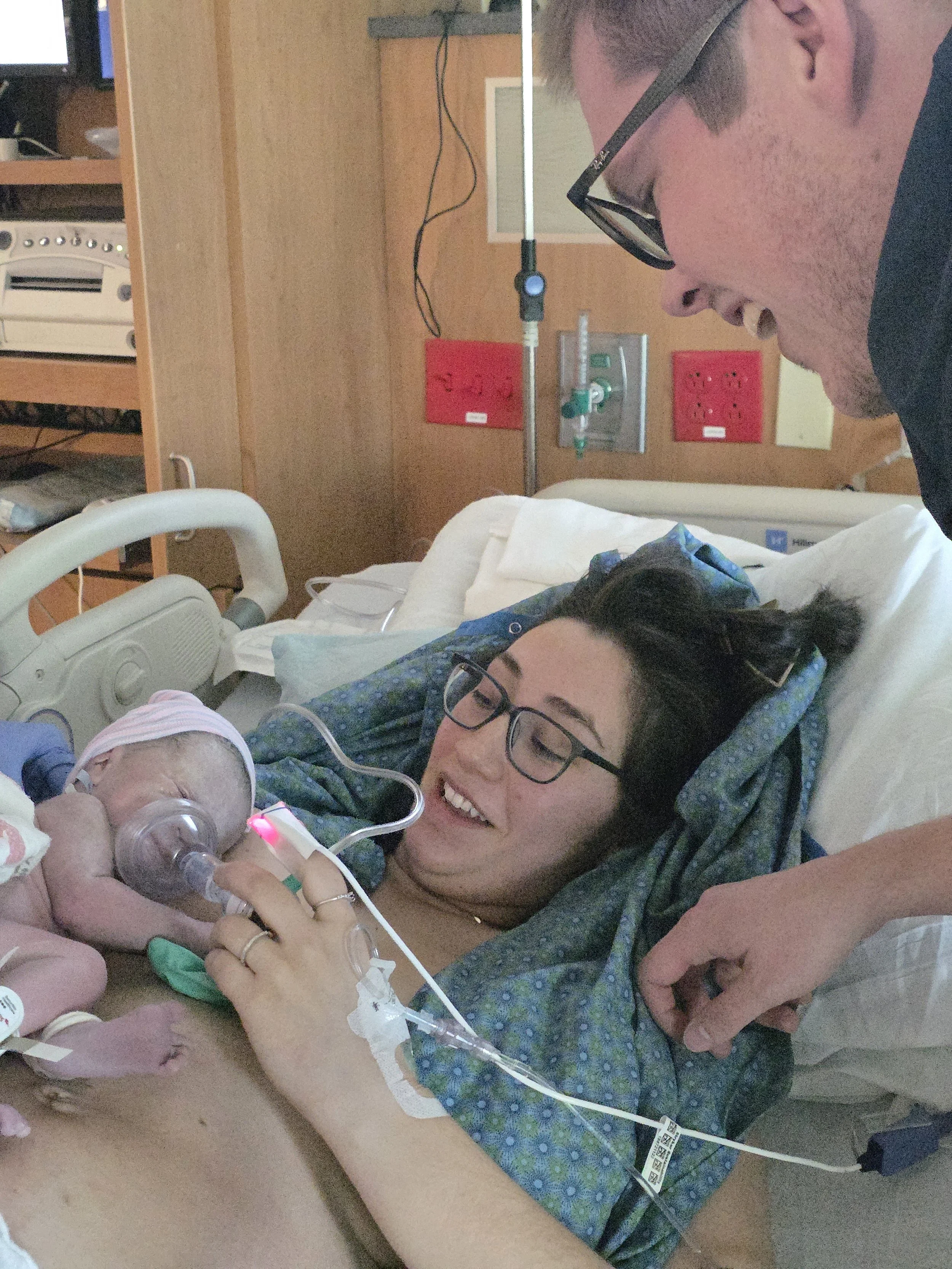 A woman lying in a hospital bed holding a newborn baby with a medical tube and oxygen mask, smiling and looking at the baby.