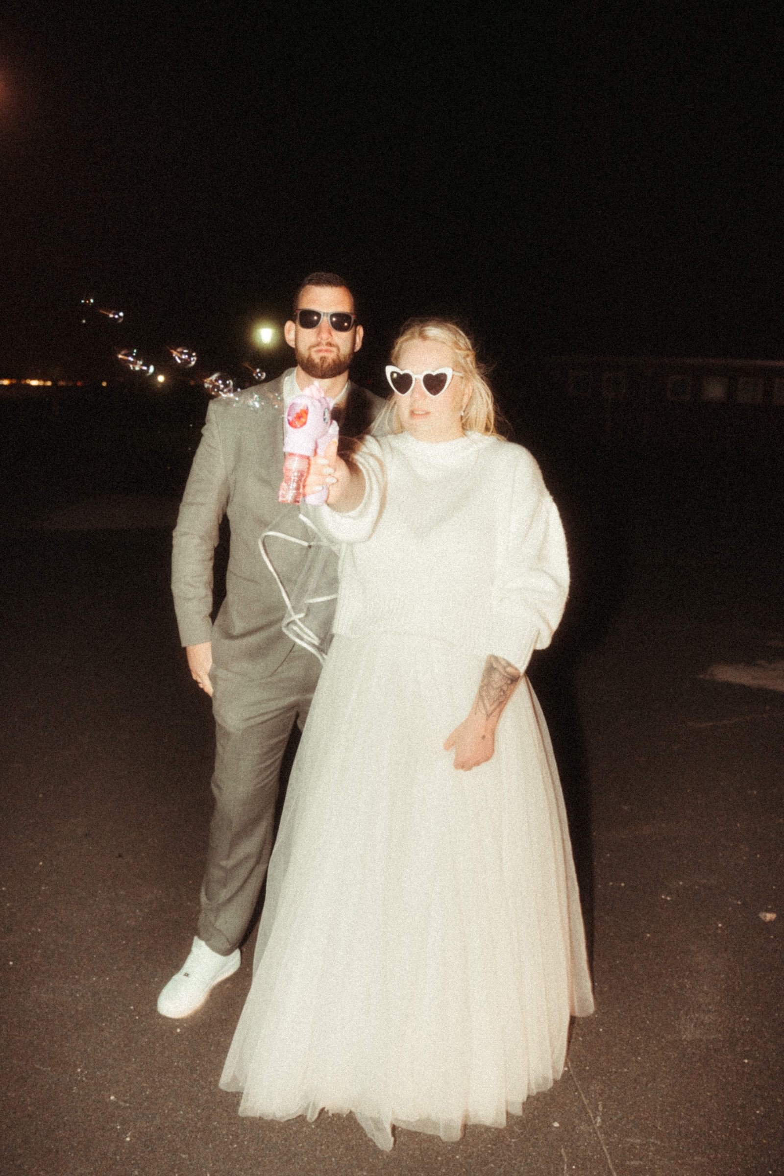 Shooting in Schillig. Ein Mann und eine Frau stehen bei Nacht auf einem dunklen Strand. Der Mann trägt einen Anzug und Sonnenbrille, die Frau trägt ein langes, weißes Kleid und Herz-Sonnenbrille, beide halten ein Buntelement in der Hand.