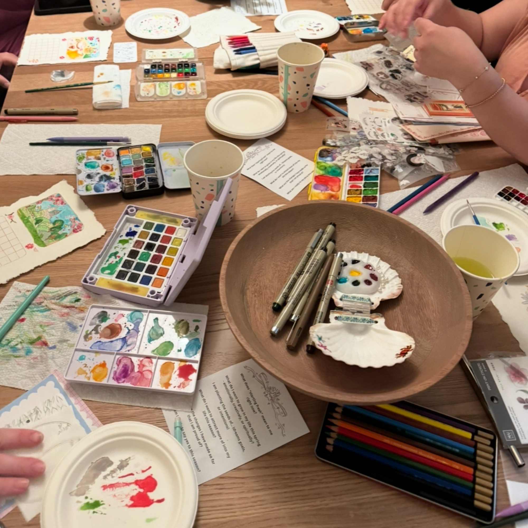 Photograph of a table clutter with pens, paints, and other art supplies.