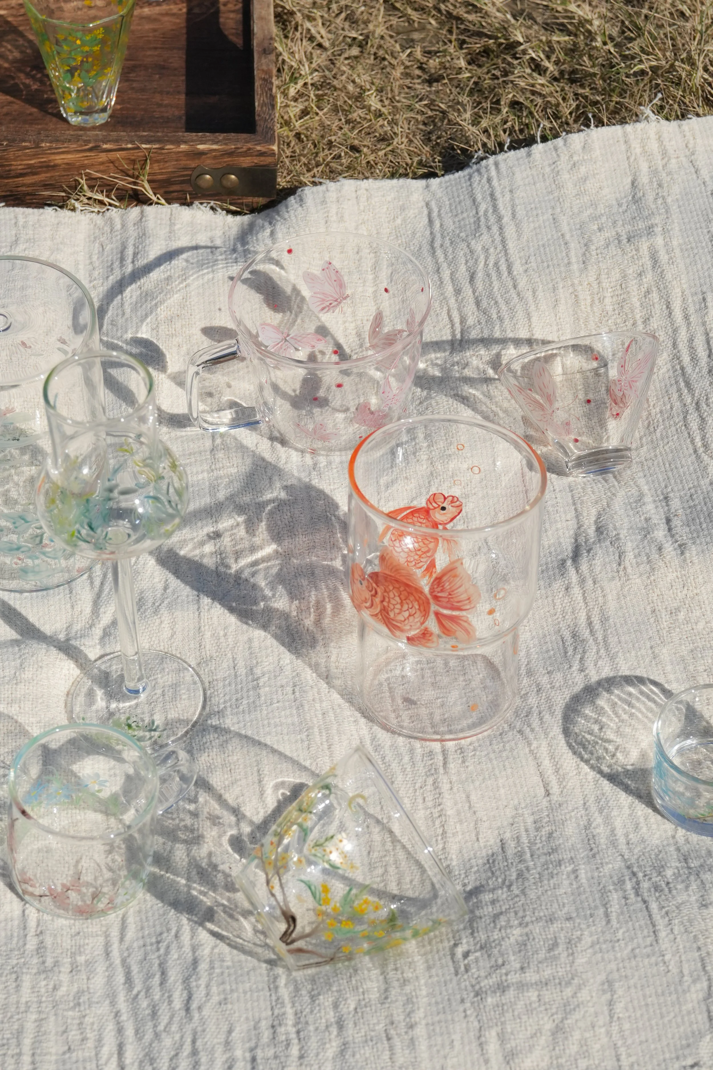 A close-up photograph of several hand-painted glass cups laying on a white blanket in the daylight.