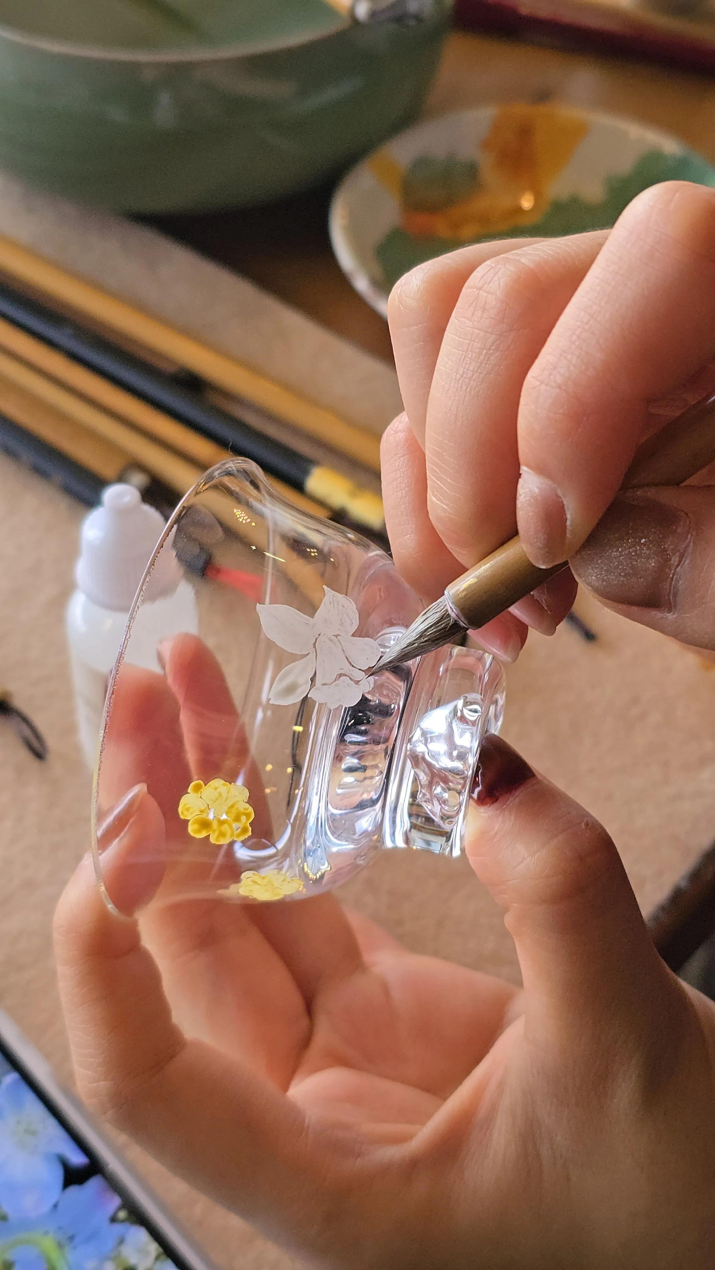 A photograph of someone gently painting a white flower onto a small glass cup. The person is holding the cup in one hand and applying paint with a brush in the other.