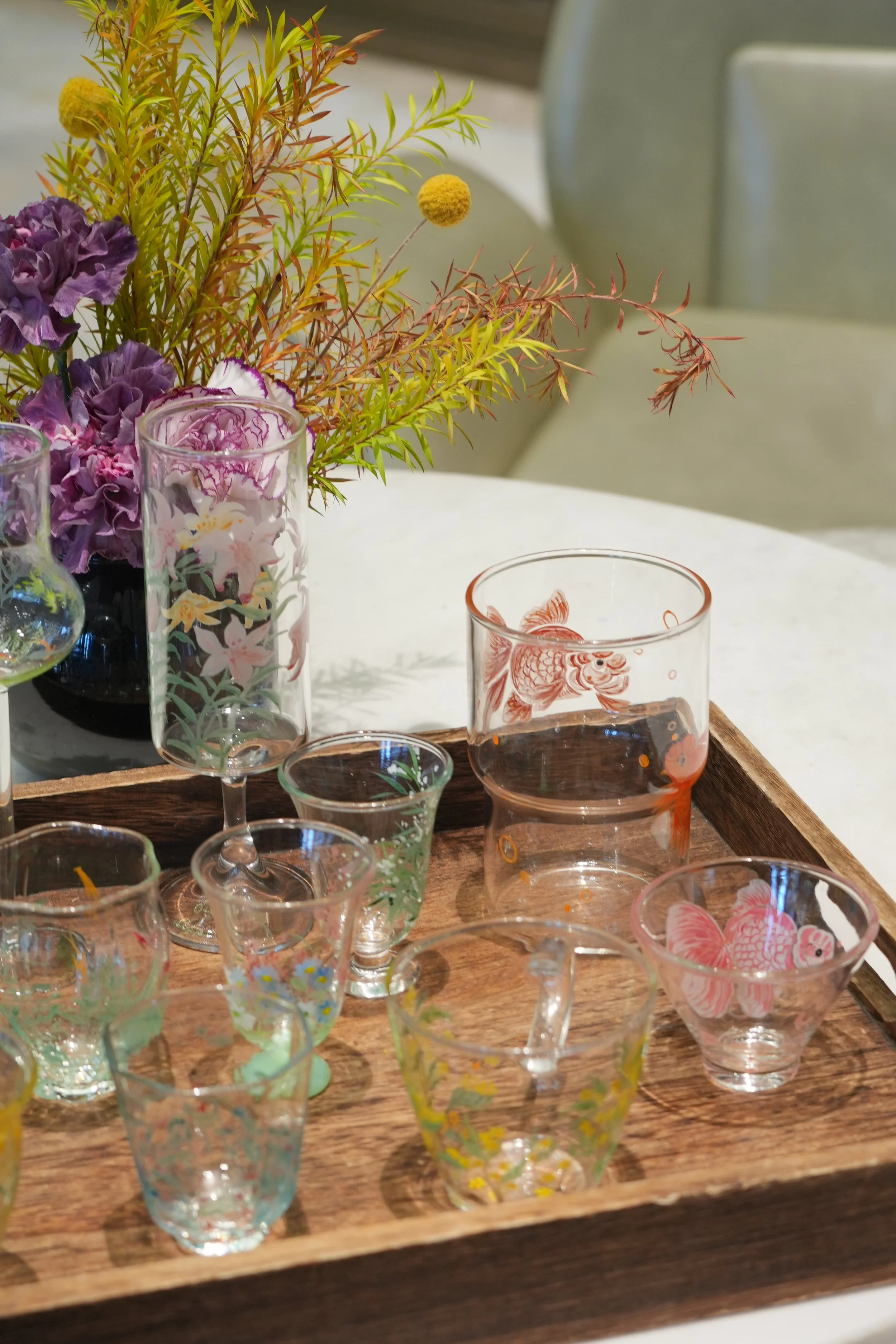 A photo of hand-painted glassware set up on a wooden tray in front of purple and green decorative flora. The glassware varies in size, height, and decoration.