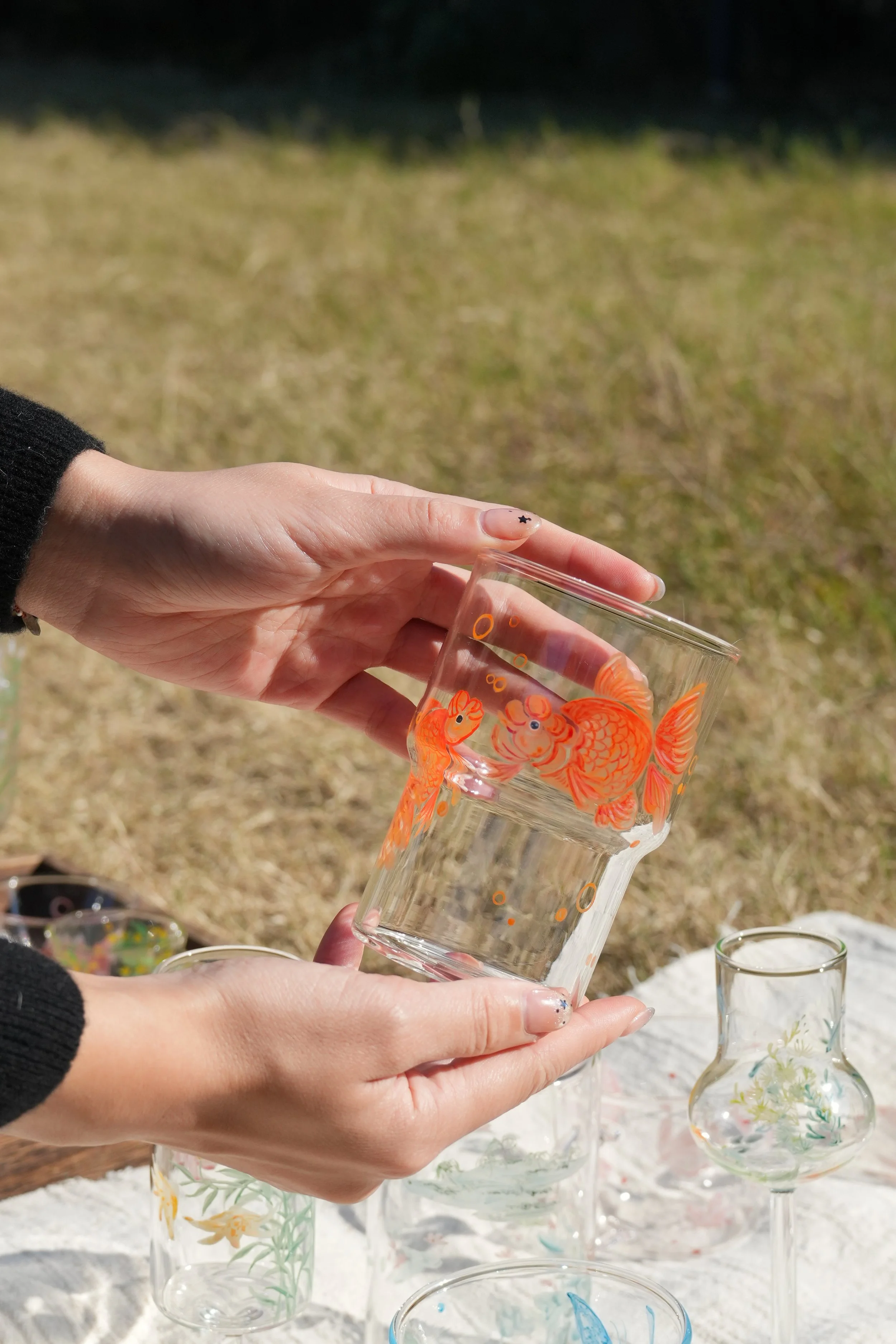 Two hands picking up a glass cup in a field. The cup is delicately painted, showing two bright orange goldfish.