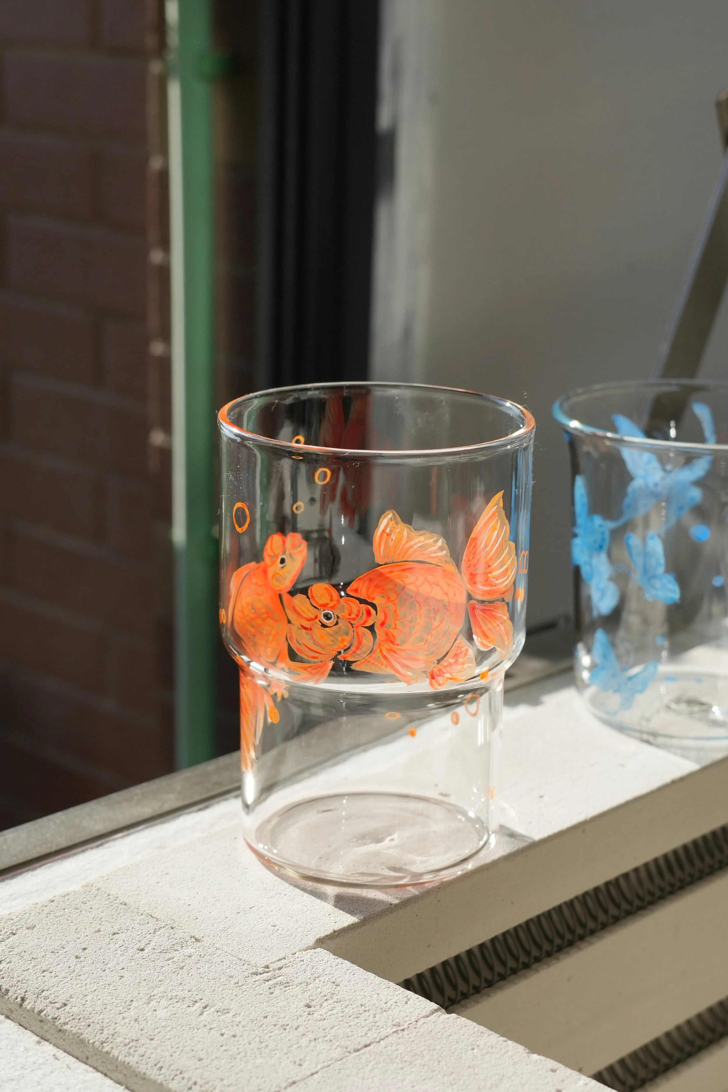 A daylight photograph of two painted glass cups: one with bright orange goldfish in the front, and another in the back with blue butterflies. The one in the back is slightly blurry,