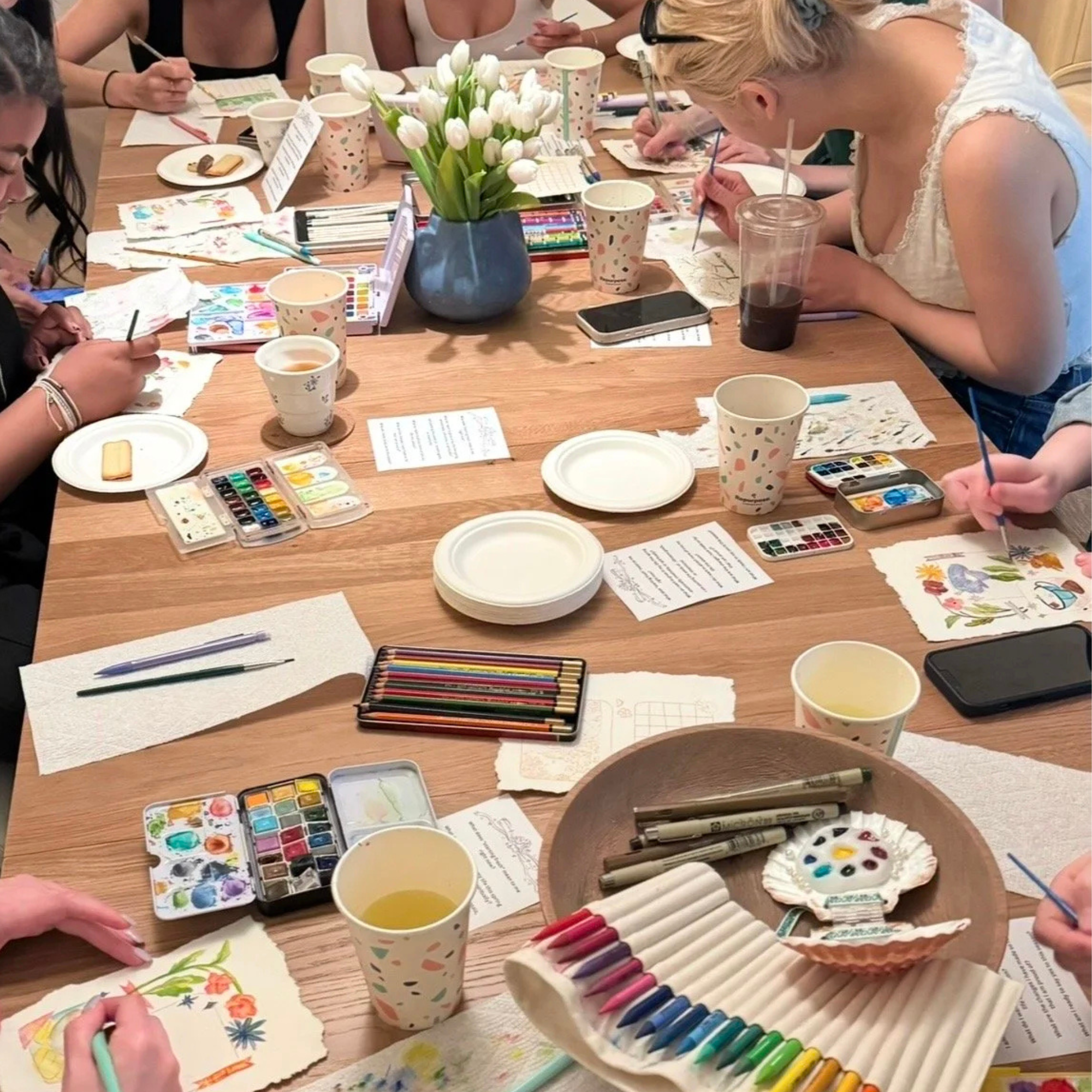 A photo of a table with people around it. On the table is flowers, craft supplies, and paint.