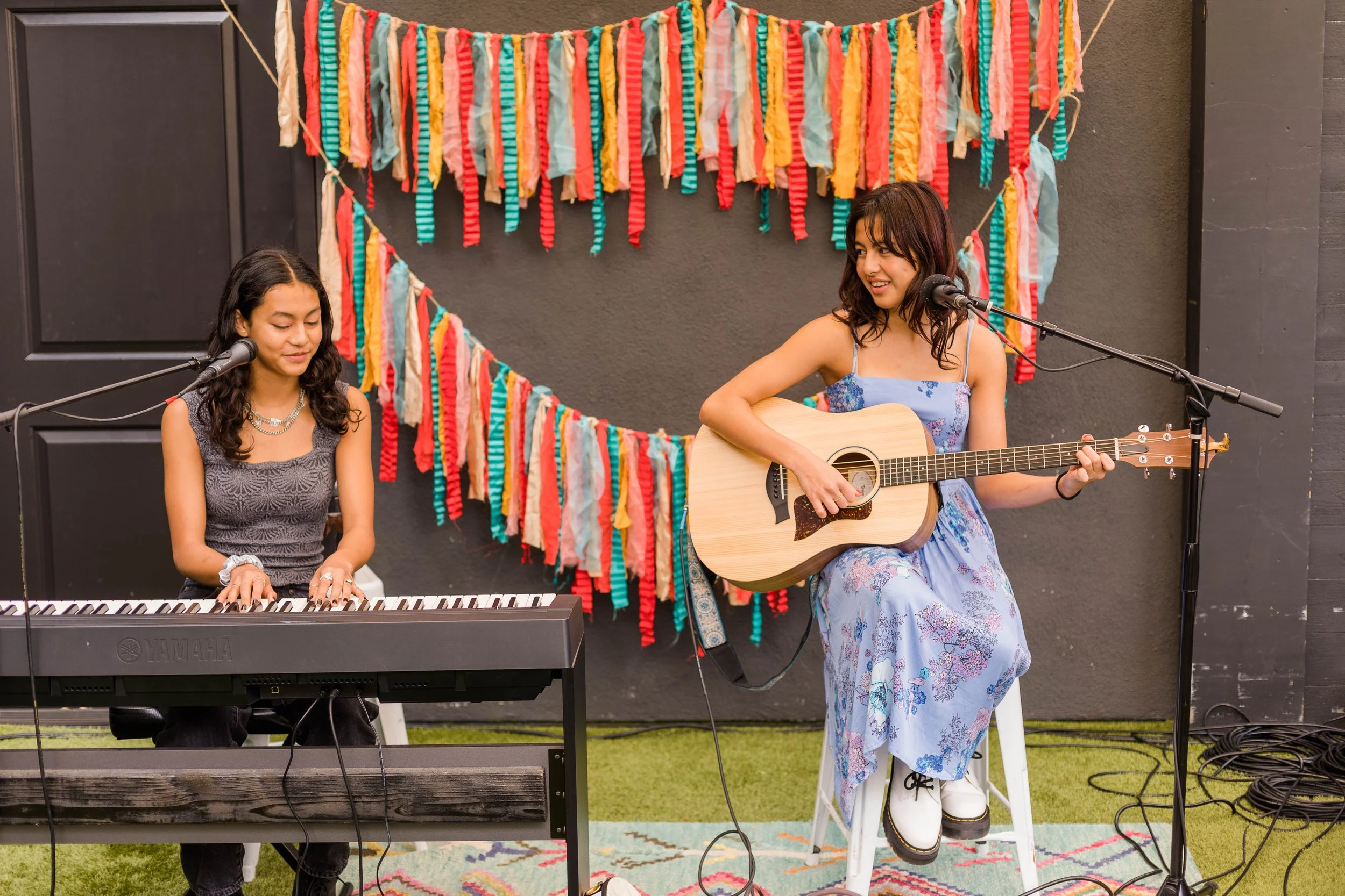 Two women perform musical act with musical instruments, one playing piano and the other guitar, in front of colorful paper garland decoration.