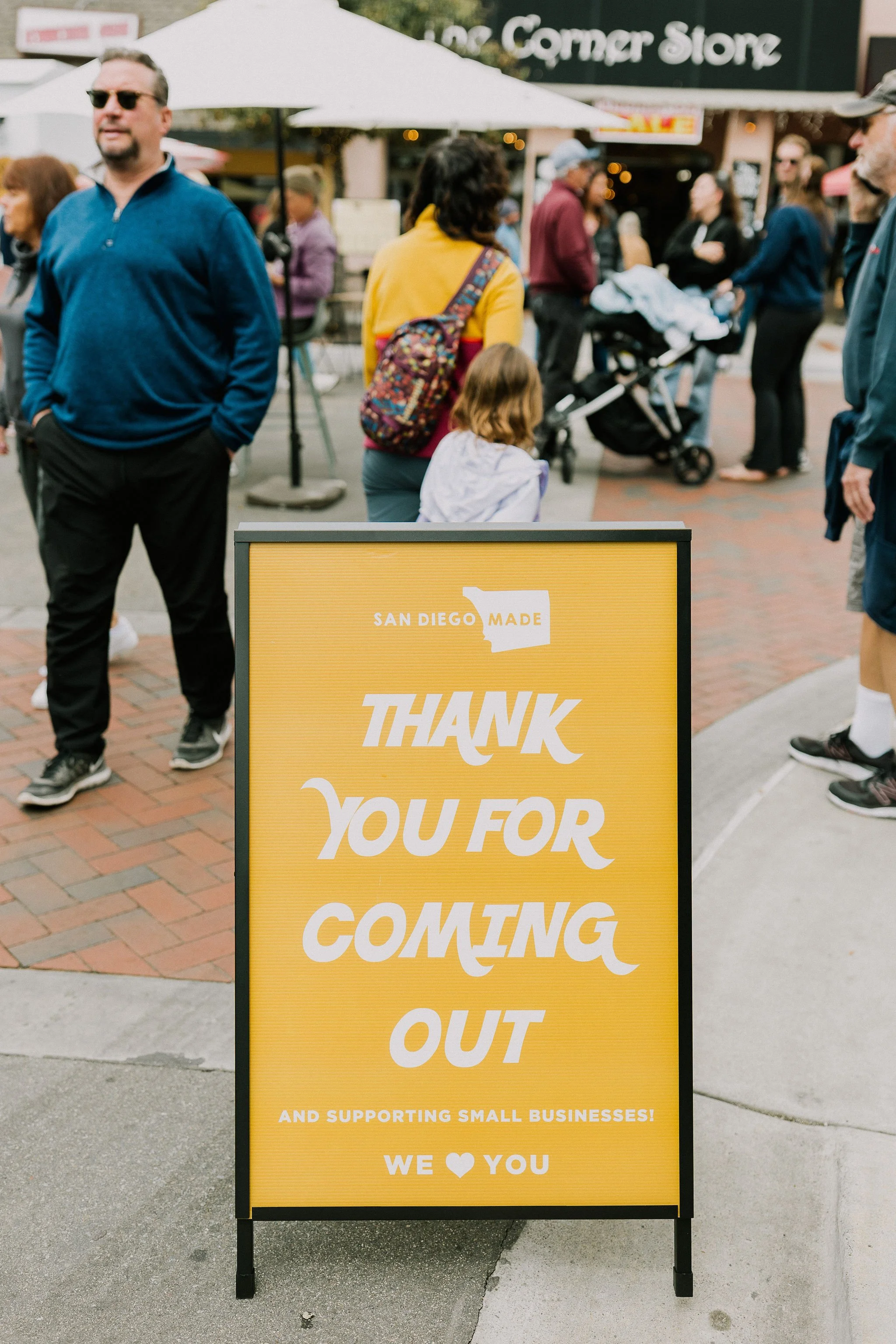 Yellow sign with white text thanking visitors for coming out and supporting small businesses, located on a brick sidewalk outside a storefront with people gathered around.