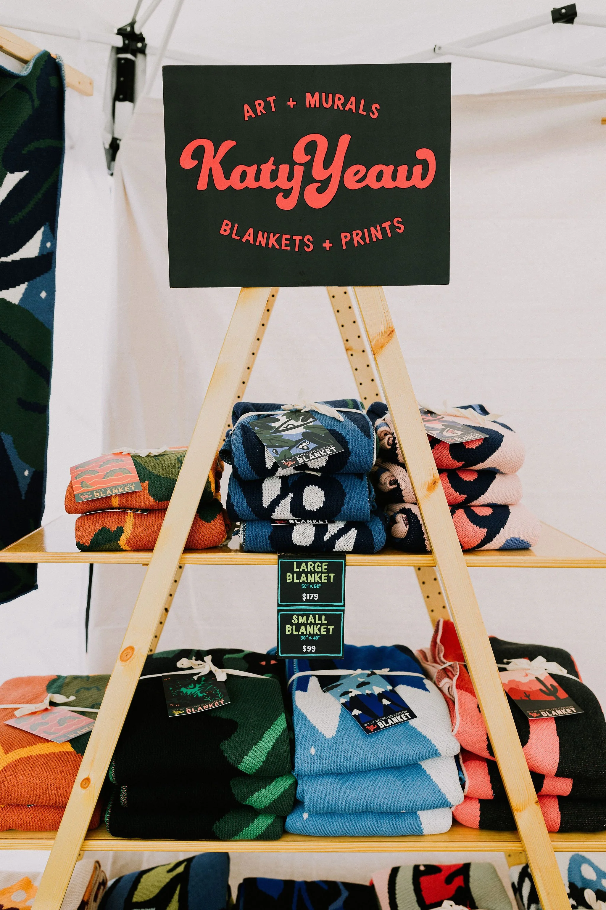 Display of colorful patterned blankets folded and stacked on wooden shelves at an craft fair, with a sign showing prices for large and small blankets.
