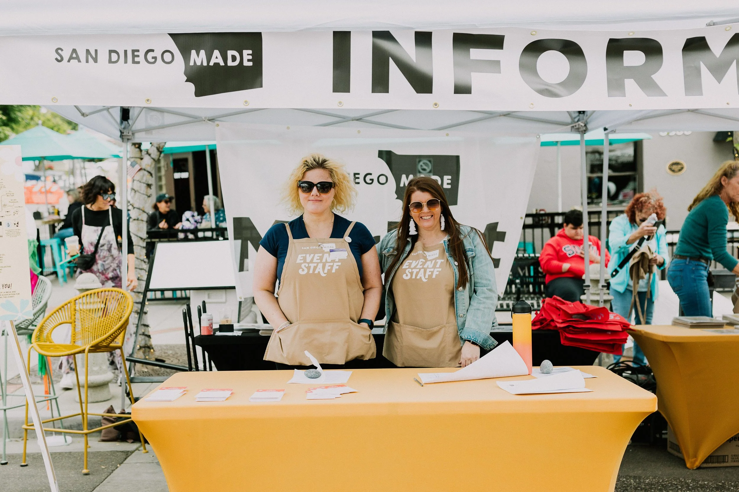 Two women in tan 'Event Staff' aprons standing behind an information booth at an outdoor event.