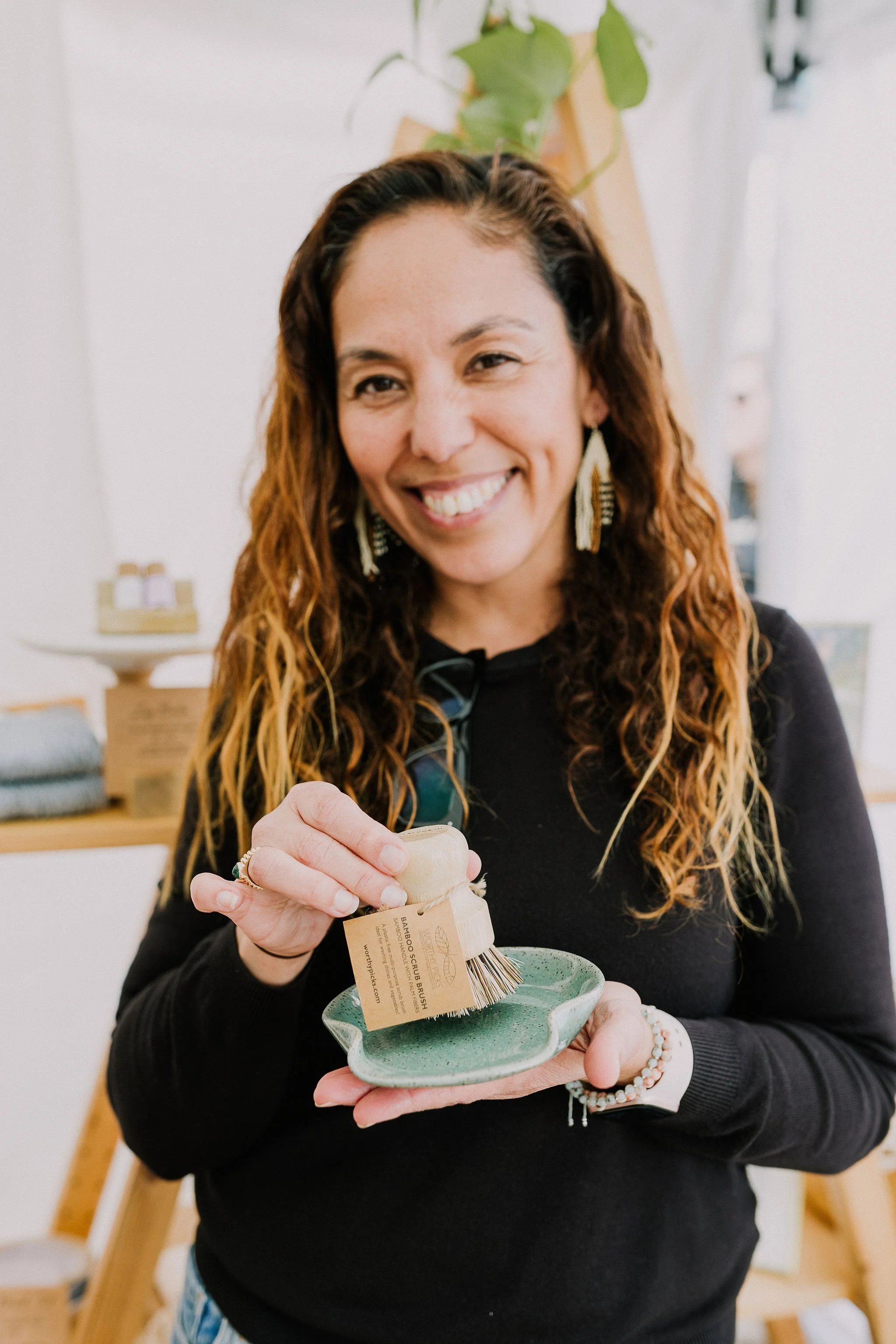 Woman with long curly hair and earrings smiling and holding a brush with a tag that reads "Bamboo Scrub Brush," standing in a well-lit room with shelves and plants.