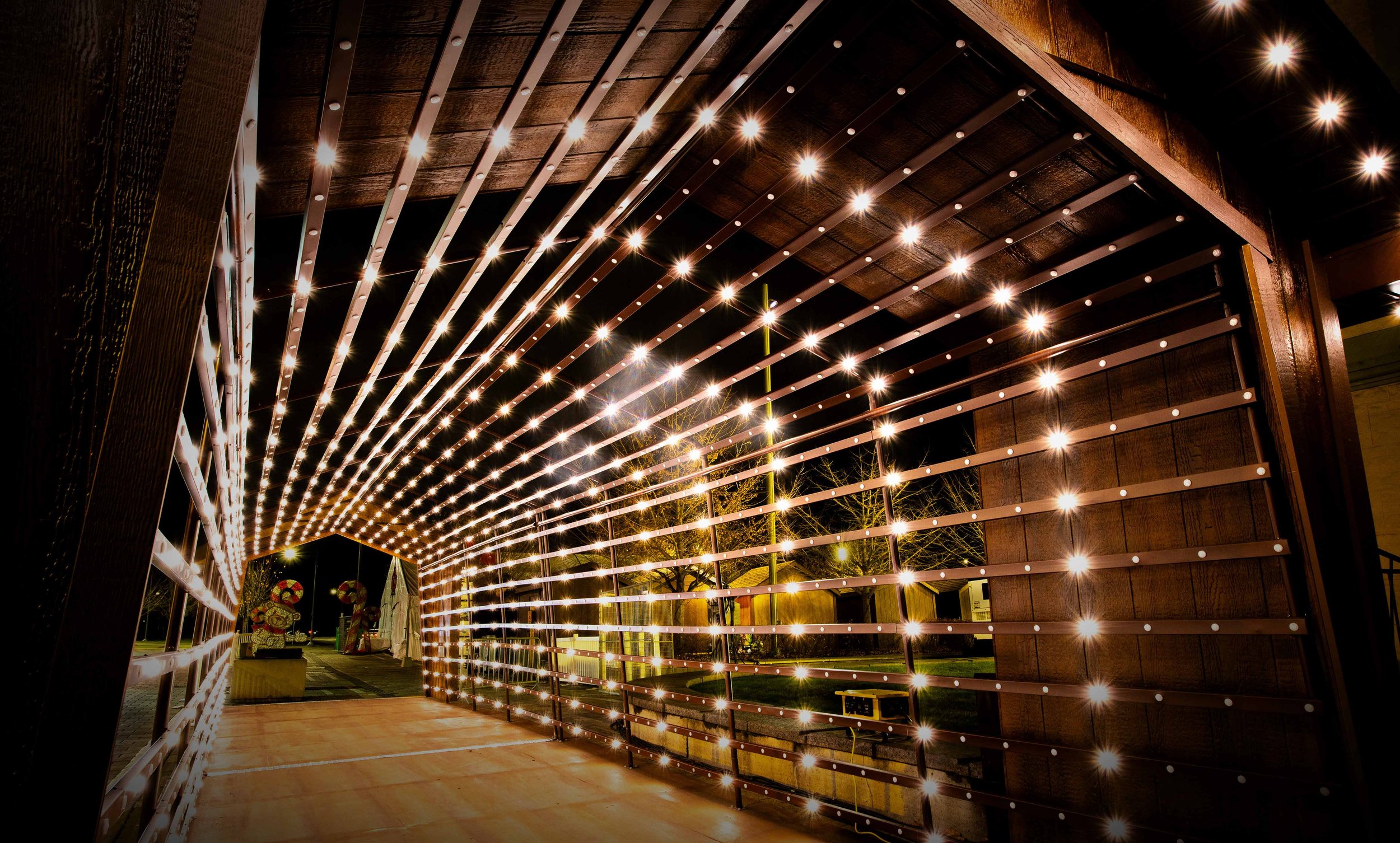 Night view of a tunnel-like structure made of dark wood with horizontal wooden slats and small bright lights illuminating the interior, with outdoor trees and streetlights visible in the background.